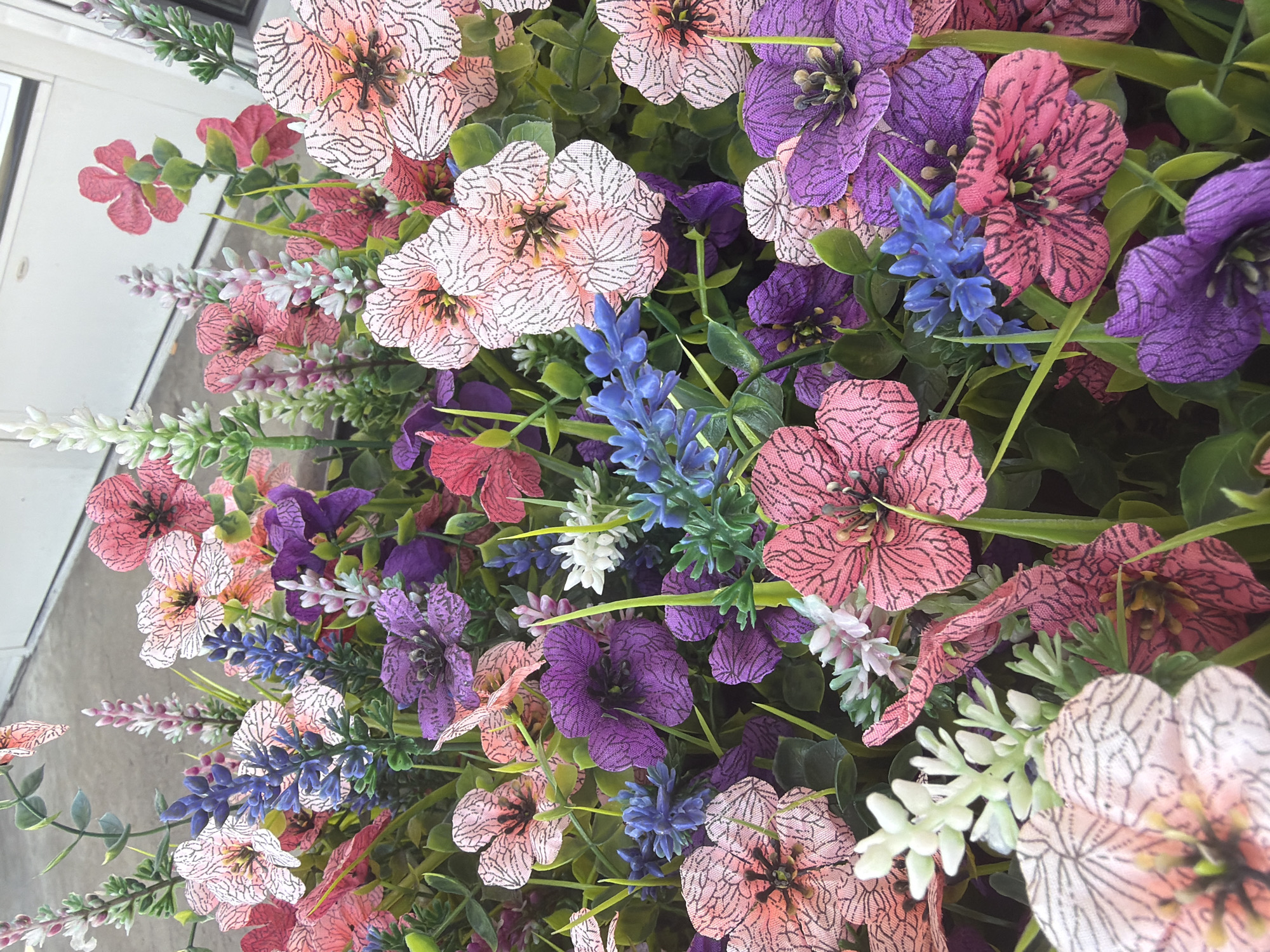 The image displays a close-up view of a vibrant floral arrangement, likely artificial due to the uniform texture and crisp detail of the petals. The arrangement is dense, with various flowers in shades of pink, purple, blue, and white, interspersed with green foliage. The pink flowers exhibit a delicate pattern of black lines on their petals, while the purple flowers are deep and velvety. Small clusters of blue and white flowers add texture and contrast. The background is blurred, suggesting a shallow depth of field, with hints of a light-colored, possibly building facade and a vertical line that could be a door or window frame. The lighting is natural and even, casting no harsh shadows. There are no people, animals, or discernible human activity in the image. The setting does not provide any specific clues about its location within the city of Lebanon, United States of America, or any urban environment.
