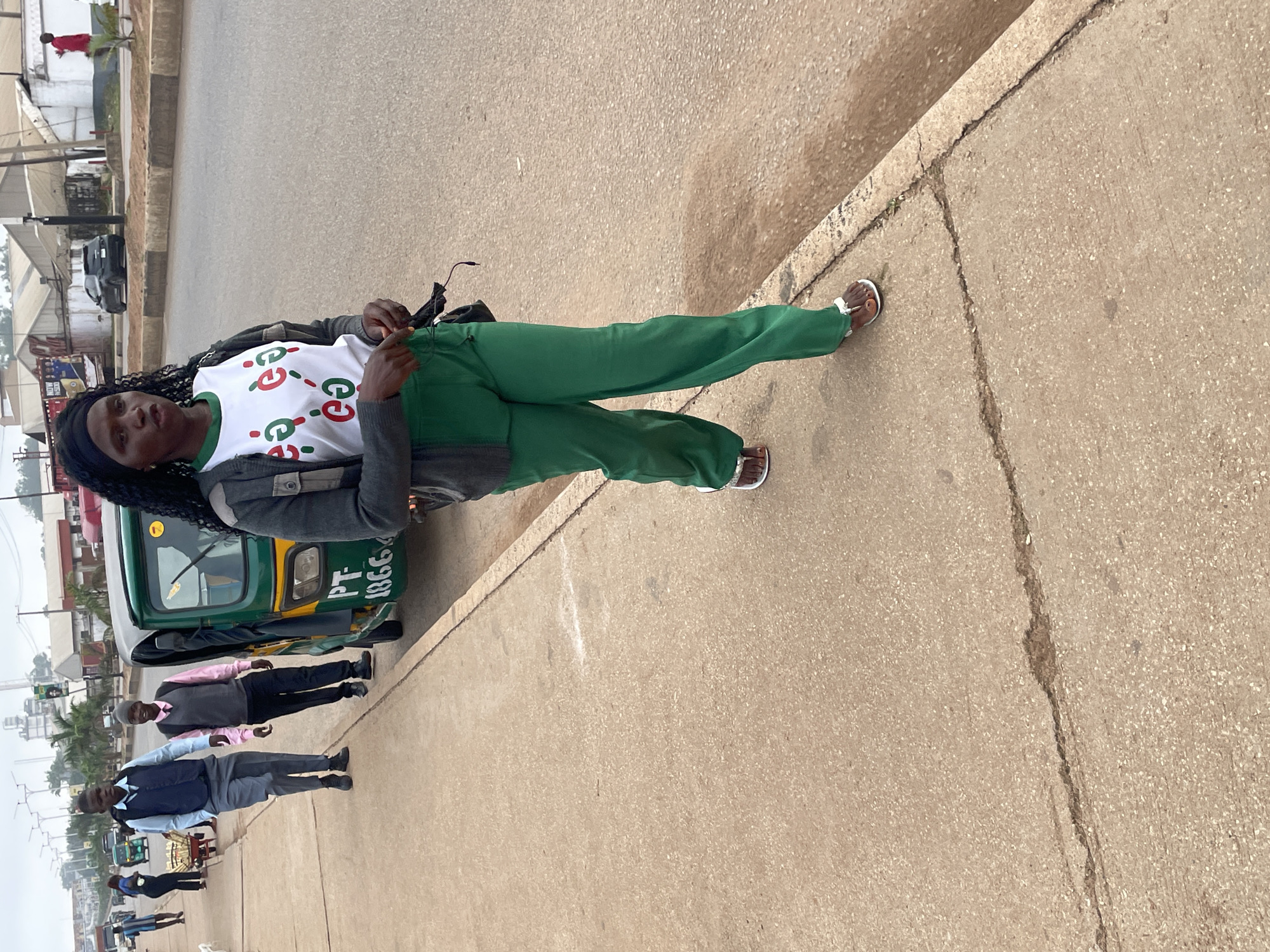 The image captures a street scene in Jos, Nigeria. In the foreground, a woman walks on a concrete sidewalk, wearing a grey cardigan over a white t-shirt with red and green circular designs, and vibrant green wide-legged trousers. She has dark, curly hair and is wearing brown sandals. To her left, a green and yellow auto-rickshaw is parked on the side of the road, with visible markings "PT 1866". Behind the auto-rickshaw, two young men are walking on the sidewalk. The man closer to the auto-rickshaw wears a light pink shirt and a grey vest, with dark trousers and a grey beanie. The man further back wears a light blue shirt, a dark blue vest, and grey trousers. They appear to be walking in the same direction as the woman. In the background, a busy street with vehicles and buildings suggests an urban environment. The sky is overcast.