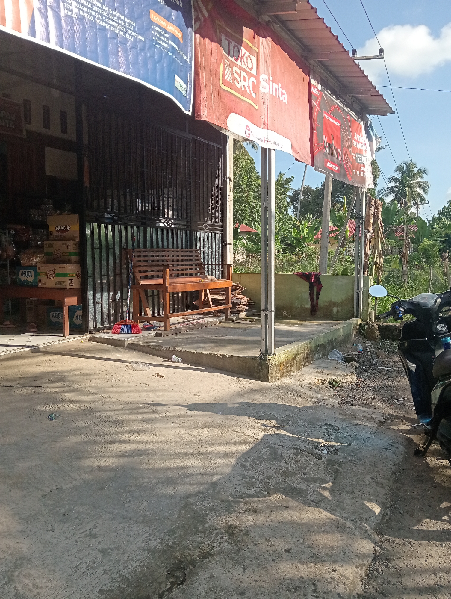 The image depicts the exterior of a small shop in Campago, Indonesia, on a sunny day. A red and white banner with "TOKO SRC Sinta" prominently displayed hangs from the storefront, along with another blue banner. In front of the shop, a wooden bench sits next to a metal gate. Stacked boxes, some with "AQUA" branding, are visible to the left. A red, blue, and yellow broom rests against the bench. A motorcycle is parked to the right of the frame, partially obscuring a concrete walkway. In the background, lush green foliage and palm trees suggest a tropical environment. The ground in the foreground is paved with concrete, showing some cracks and debris. A red cloth is hanging on a wall in the middle ground.