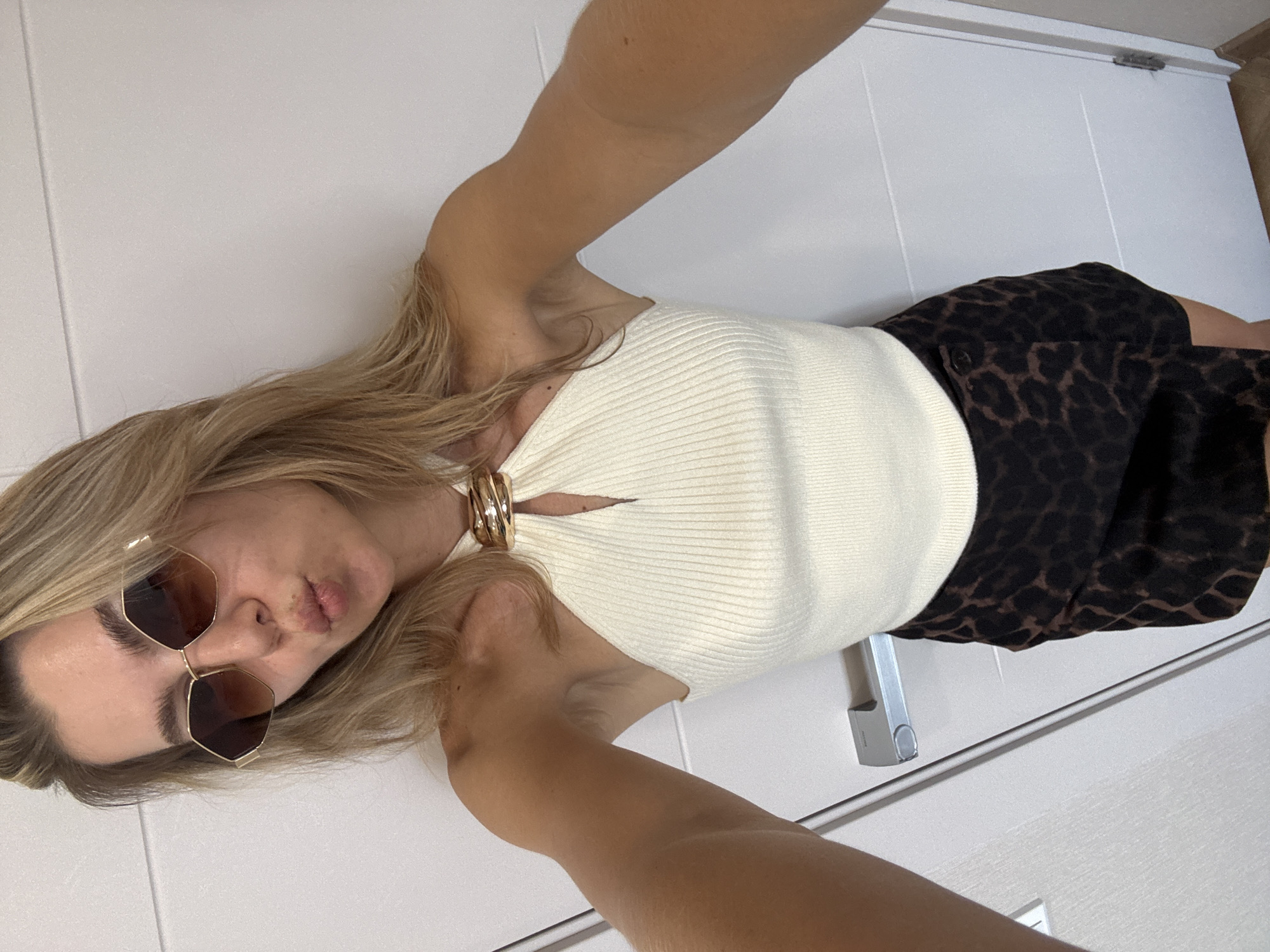 A young woman with blonde hair and sunglasses poses for a selfie against a white door. She is wearing a cream-colored halter top with a gold accent at the neckline and a dark leopard-print skirt. Her lips are puckered.
