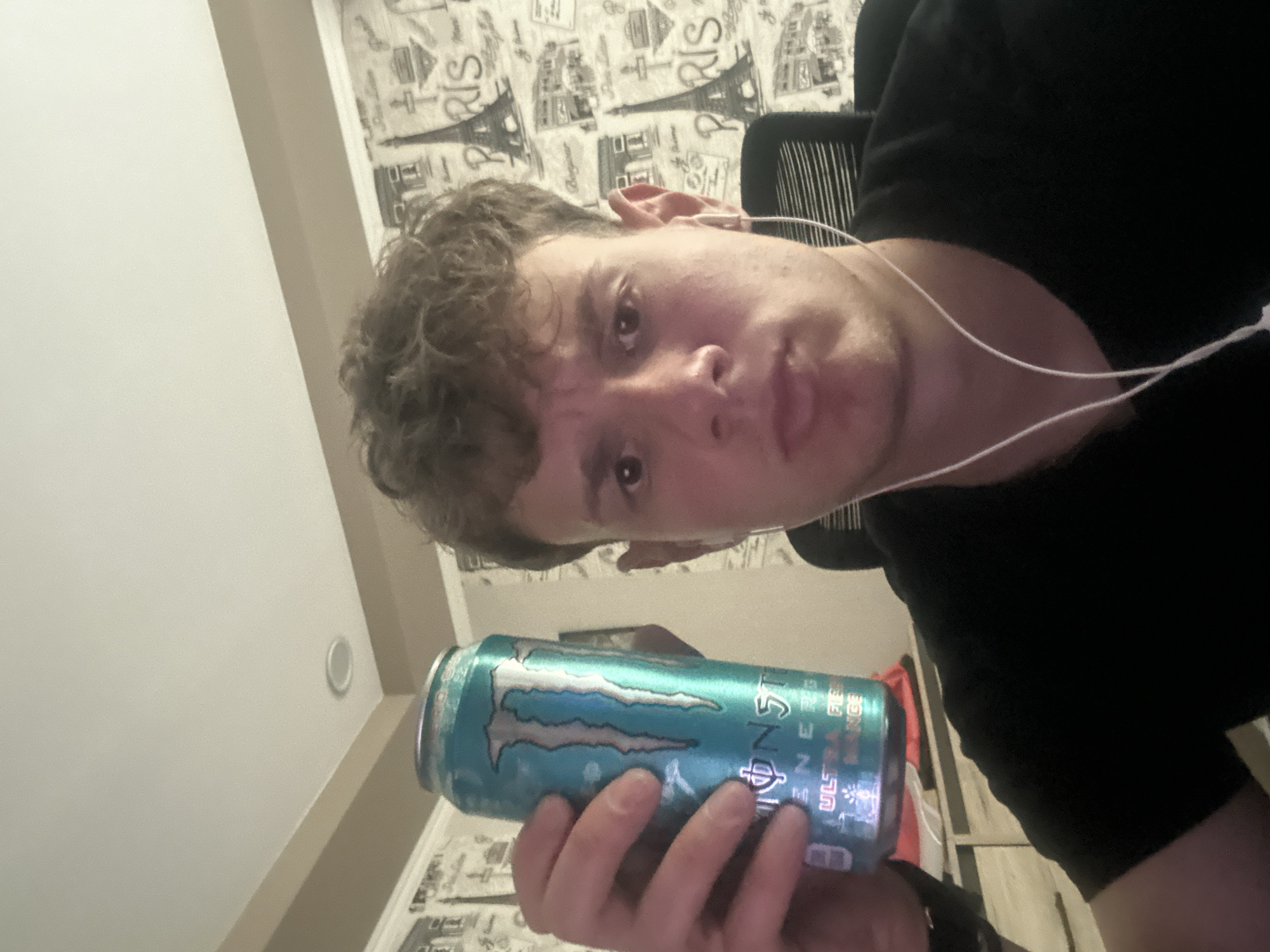 A man is holding a can of Monster Energy Ultra Fiesta Mango in his left hand. He is wearing earbuds and a black t-shirt. He is sitting in a chair with a black mesh back. The wall behind him has wallpaper with a pattern of Eiffel Towers and the word "Paris". The location is likely indoors. The location in the city of Elhovo, Bulgaria is not verifiable from the image.