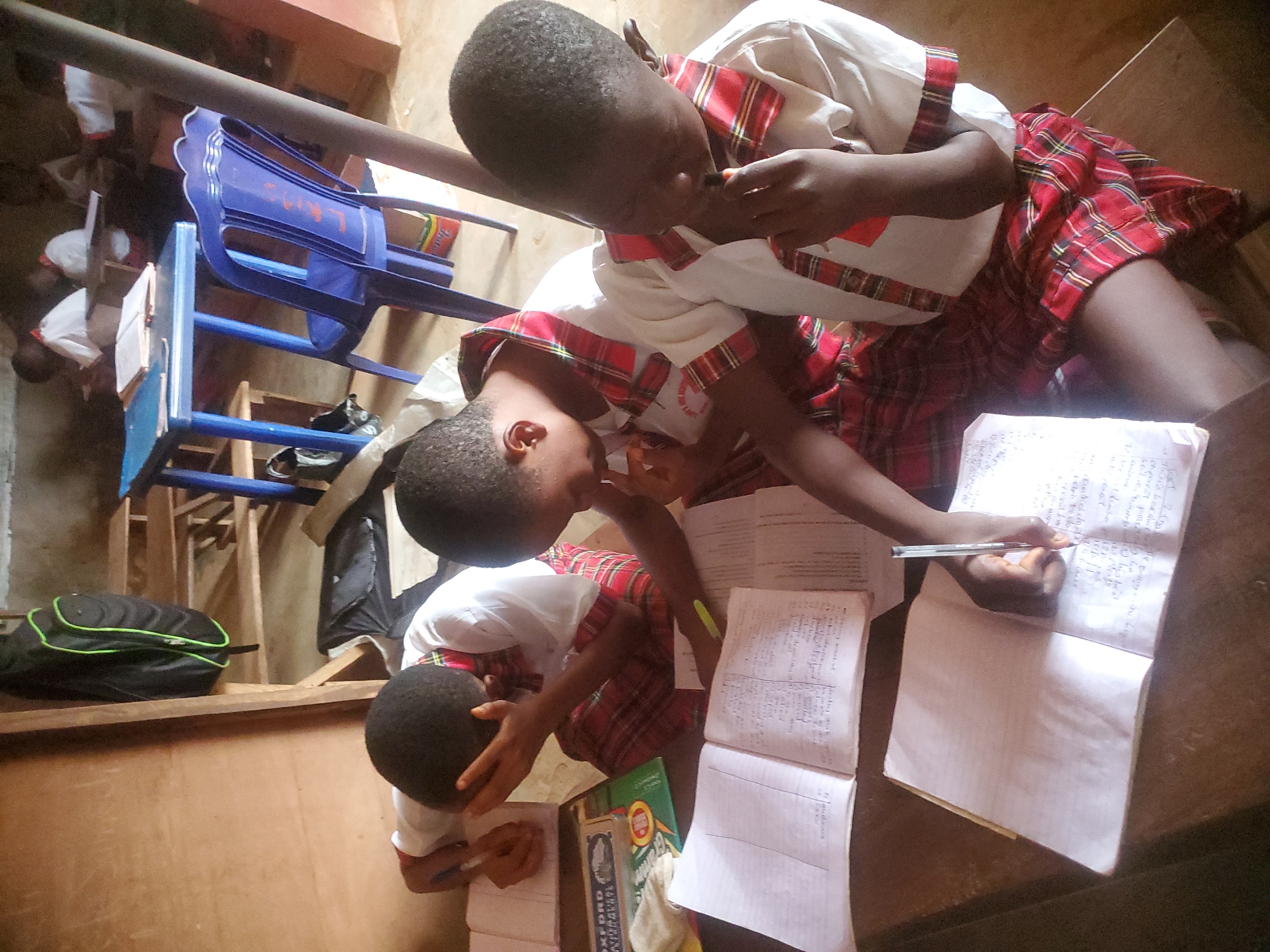 In Ukwu-Abwa, Nigeria, three children in school uniforms are writing in notebooks. The uniforms consist of white tops with red plaid trim and red plaid skirts. One child holds a pen and is actively writing. The children are surrounded by notebooks and textbooks. Blue plastic chairs and a black backpack with green trim are in the background. The setting appears to be indoors.