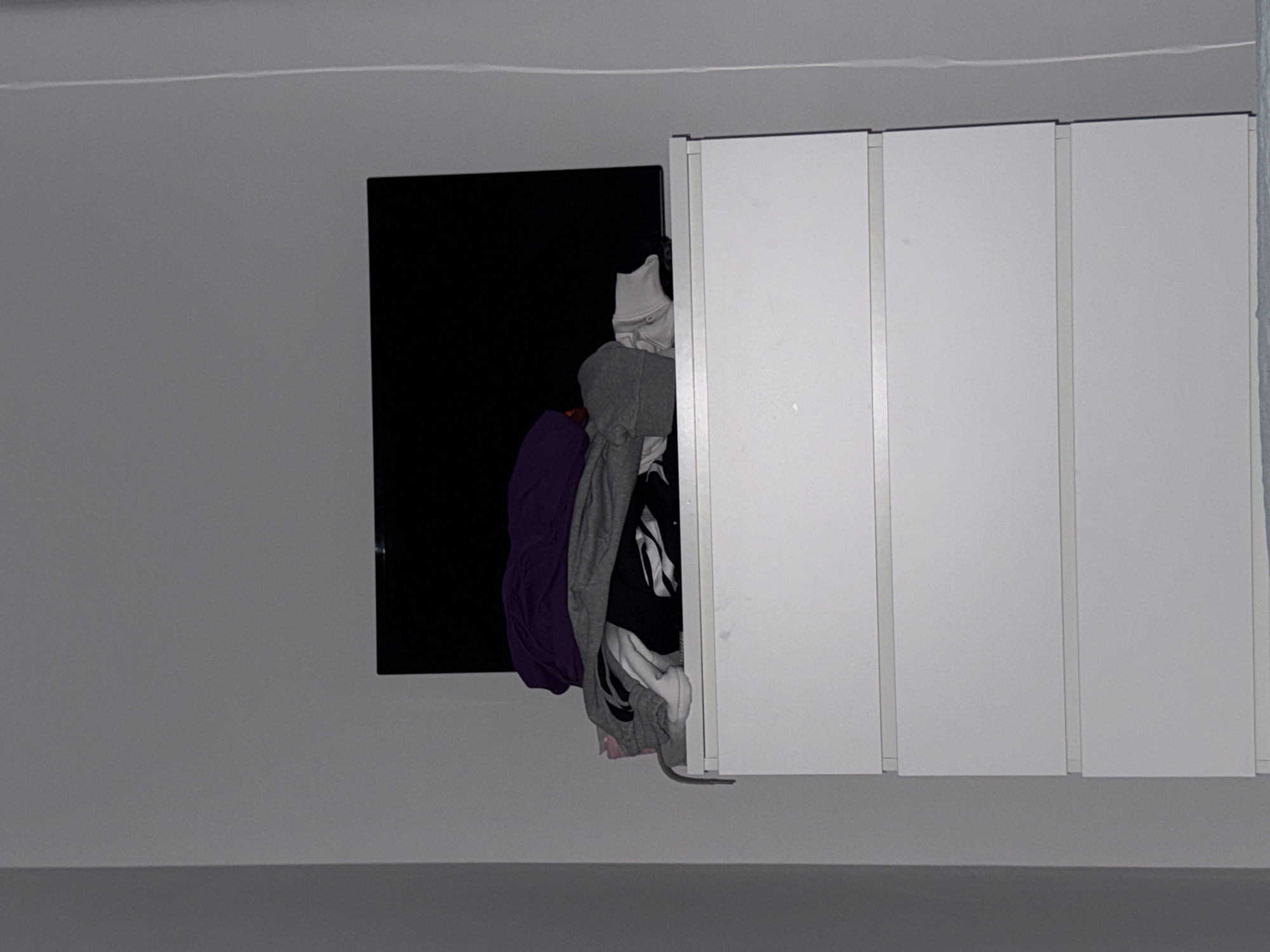 The image depicts a white wall with a partially open cabinet. The cabinet has four vertical, rectangular doors. The leftmost door is open, revealing a black interior filled with assorted clothing, including a purple shirt, a gray hooded sweatshirt, and other white and black garments. The right three doors are closed and appear white with gray seams. The scene appears to be indoors. The location in Stara Zagora, Bulgaria is not evident from the image.