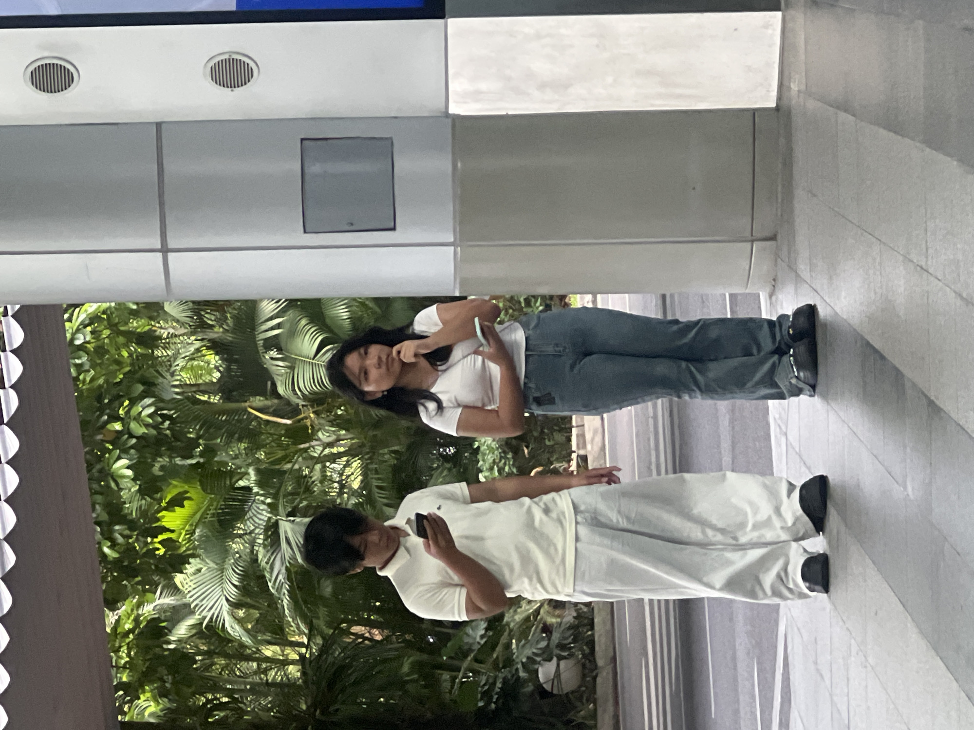Two young individuals are standing on a paved sidewalk. A male on the left, wearing a white polo shirt and loose white pants, is looking down at his smartphone. A female on the right, in a white cropped t-shirt and blue jeans, is looking to her right, with her hand resting on her chin and holding a smartphone. Behind them, lush green foliage suggests an outdoor, possibly tropical, environment. The background also features a modern building with metallic cylindrical columns and circular vents. The image appears to have been taken in an urban setting in Da Nang, Vietnam, likely in a public space like a park or along a thoroughfare with extensive landscaping.