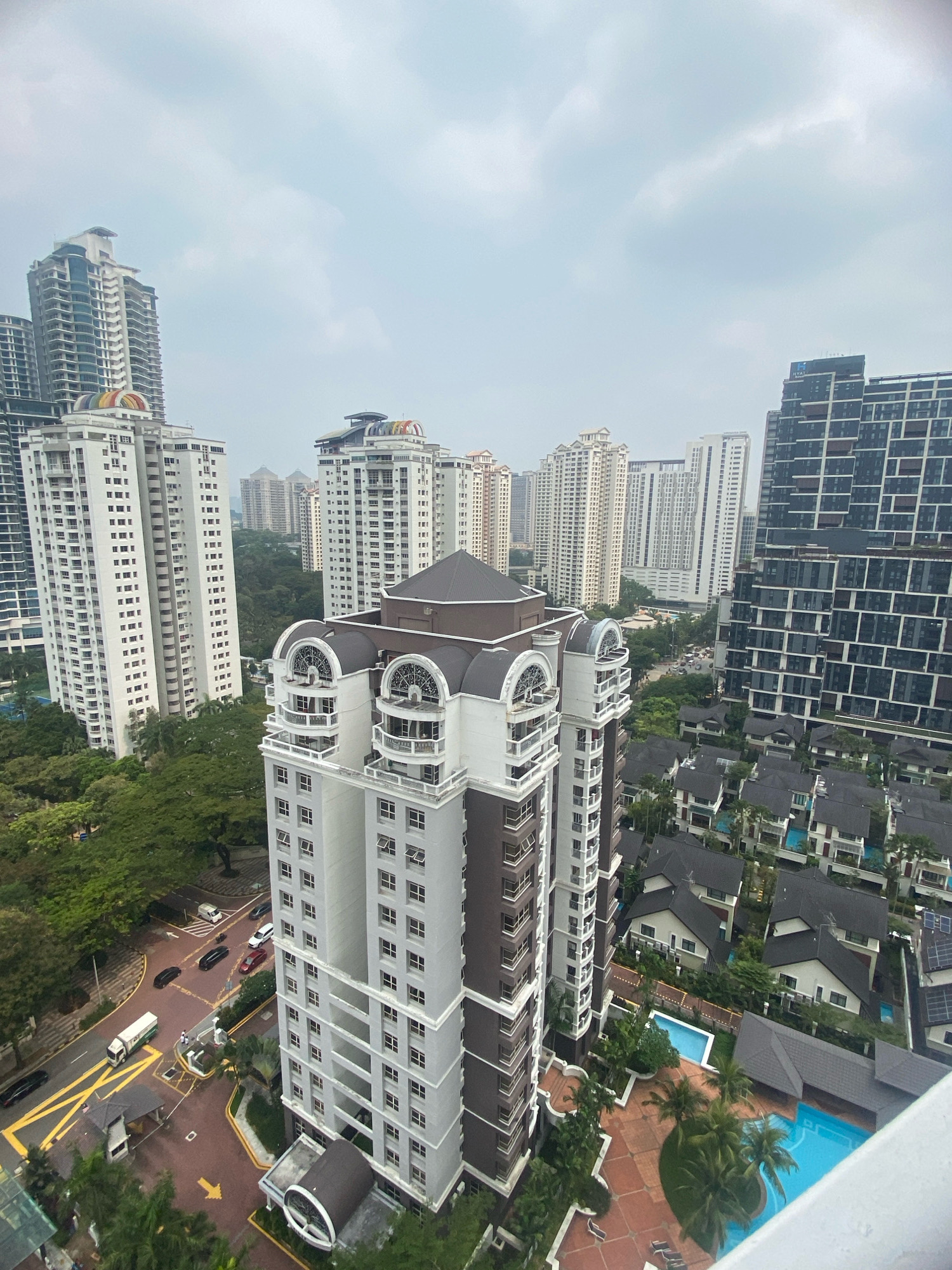 This is an aerial view of a residential area in Kuala Lumpur, Malaysia. The scene is dominated by a collection of modern high-rise apartment buildings and low-rise houses with dark roofs. In the foreground, a striking white and brown high-rise building with arched window features and balconies stands out. To its right, a darker, more modern high-rise building with a grid-like facade is visible, featuring numerous windows. In the background, a dense cluster of tall apartment buildings and some smaller residential structures can be seen, interspersed with lush green trees. A road with several cars, including a white truck and some black sedans, is present, indicating an active urban environment. The sky is overcast with light clouds. The residential area appears well-maintained, with visible swimming pools and manicured landscaping in some of the housing developments.