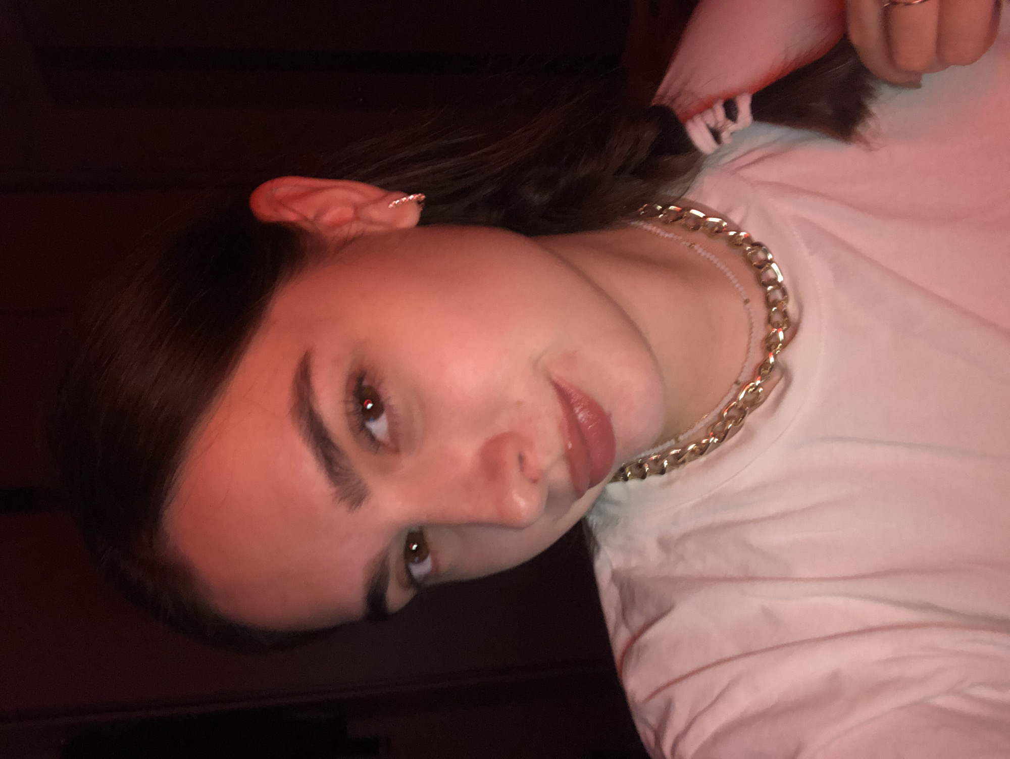 This is a close-up, self-portrait photograph of a young woman indoors, with a red light source casting a warm glow on her face and clothing. The background is dark and out of focus, suggesting an indoor environment with low lighting. The woman has brown hair, styled in a braid that is visible on her right side. She is wearing a white t-shirt and two necklaces: a thicker gold chain and a thinner, beaded necklace. She has on small hoop earrings. The lighting creates a dramatic effect, highlighting her facial features and accessories. There is no discernible information about the city of Noviy, Russia, or any specific location within it present in the image.