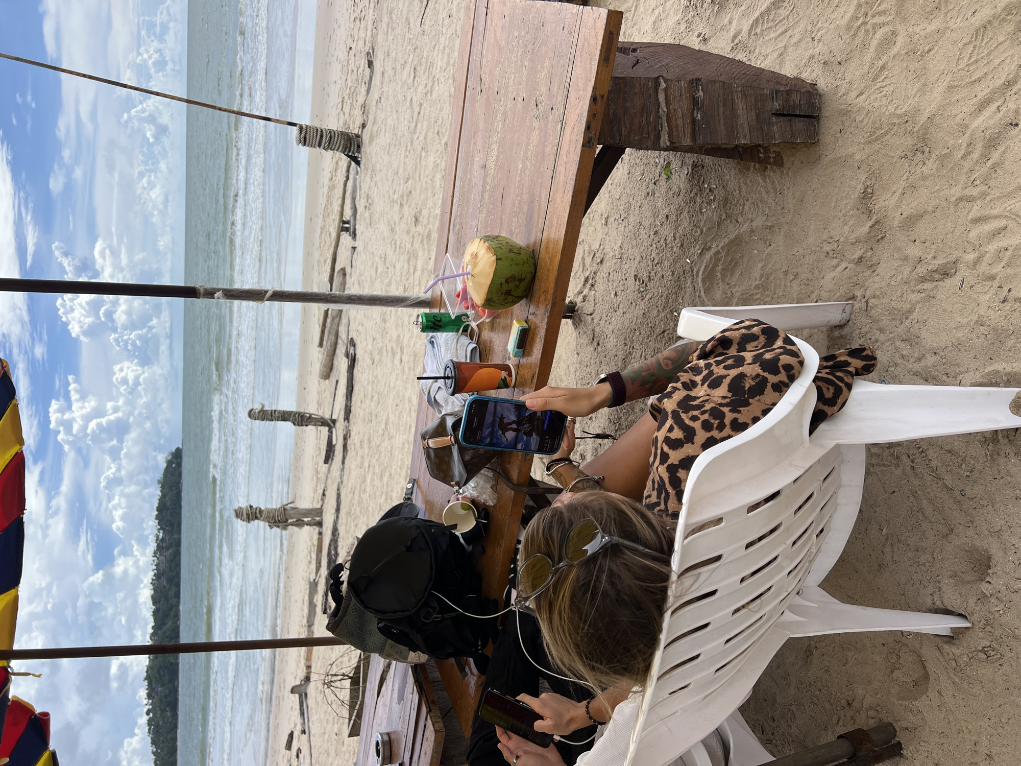 A group of people are sitting at a wooden table on a sandy beach. The table is set with drinks, including a coconut with a straw, and cans. A person in a leopard print outfit is holding a smartphone. Another person is using a smartphone and wearing sunglasses. There is a backpack on the table. In the background, the ocean and sky are visible. The weather appears to be sunny with some clouds. The overall scene suggests a relaxed beach setting.