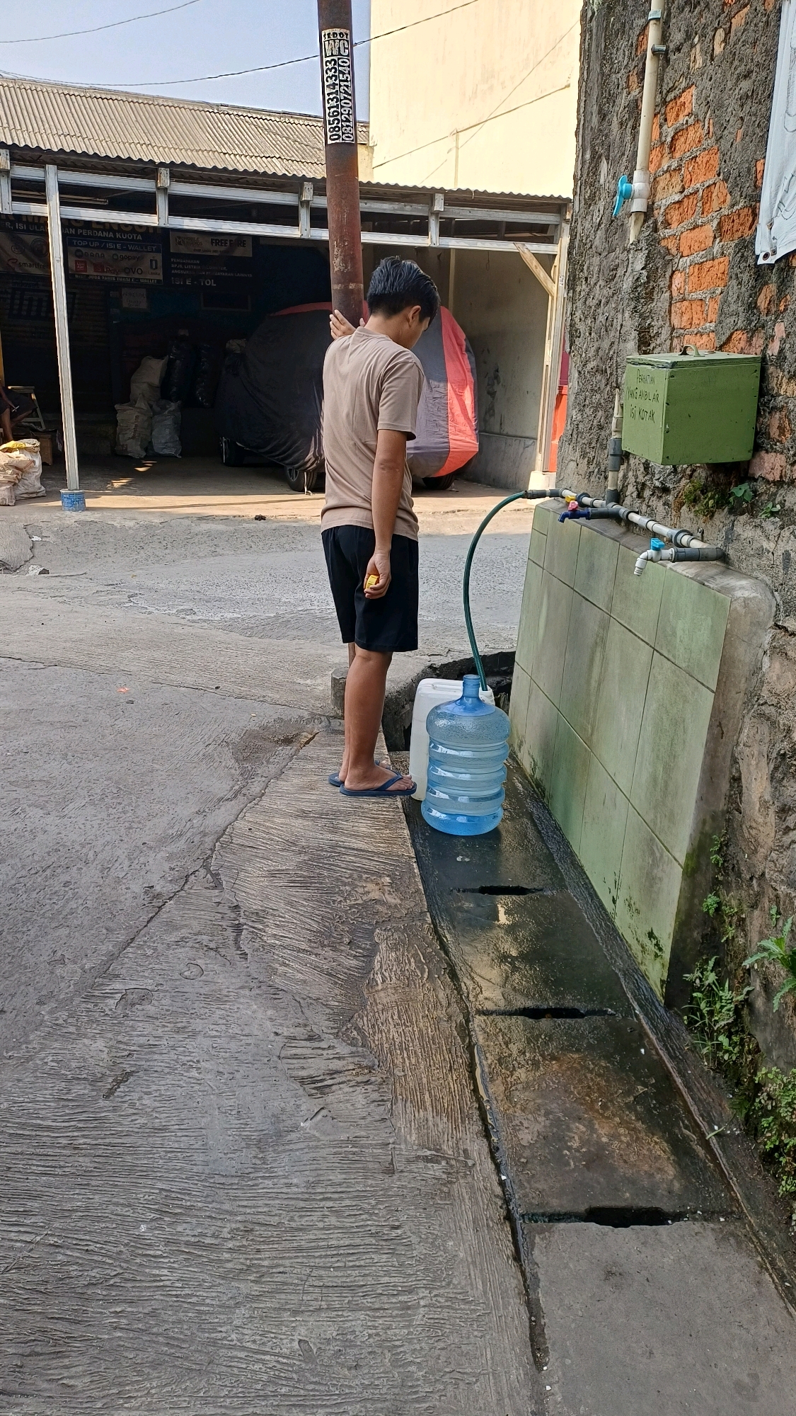 A young man wearing a light brown t-shirt and black shorts stands on a concrete sidewalk next to a drainage ditch, holding a yellow object in his right hand. He is facing a large, clear plastic water jug that is being filled by a green hose connected to a faucet on a brick wall. The jug sits on the edge of the drainage ditch, which contains some water. Behind the man is a building with a corrugated metal roof. A pole with stickers is visible in the foreground. A green box is mounted on the brick wall above the faucet, with text on it. The scene appears to be in a semi-urban environment in Citeureup, Indonesia, indicated by the signage and general architecture.