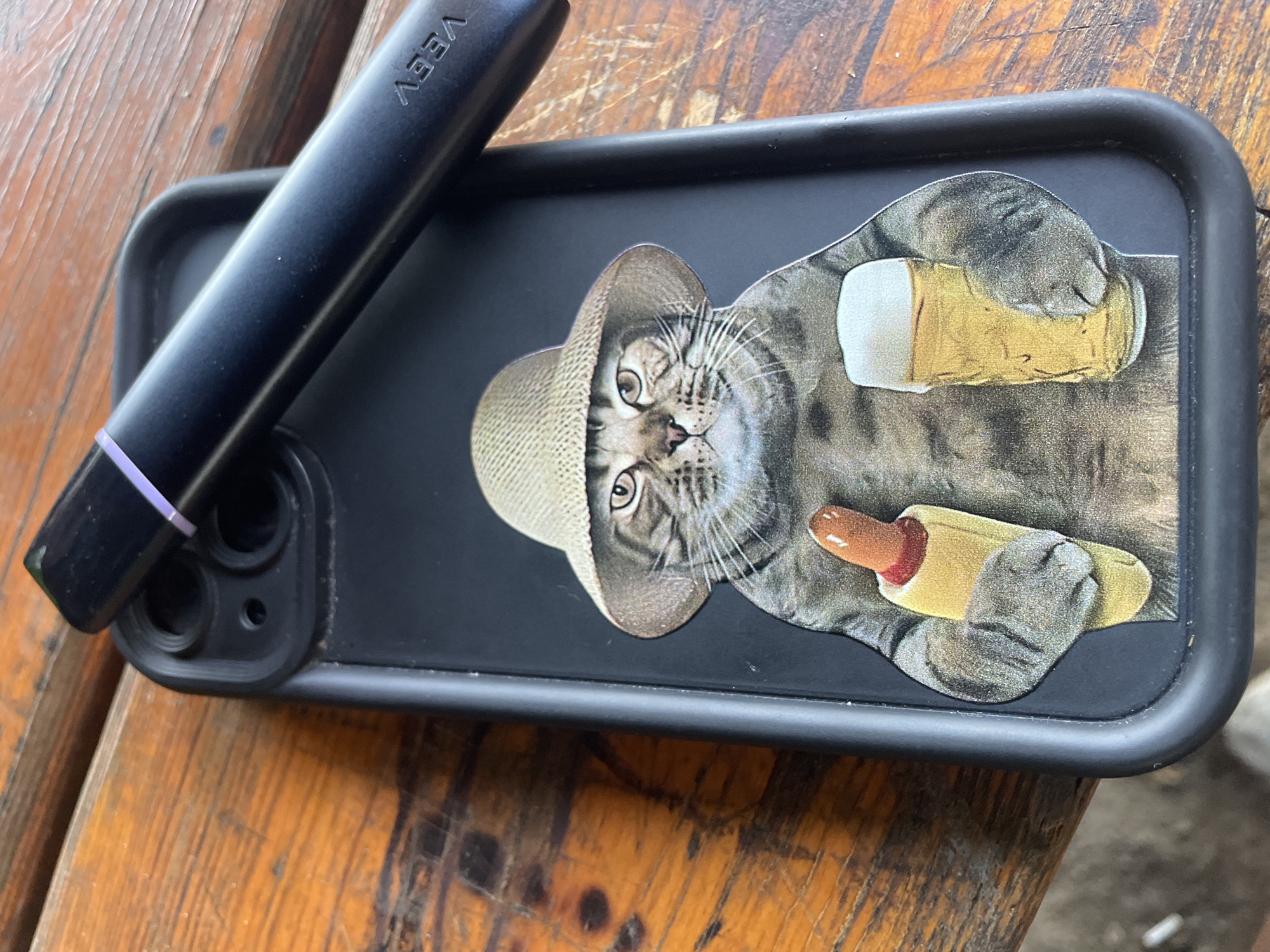 A black iPhone case with a printed image of a tabby cat wearing a straw hat and holding a hot dog in its left paw and a glass of beer in its right paw rests on a wooden surface. A black vape pen with a purple band near the mouthpiece lies diagonally across the top of the iPhone case. The wooden surface appears to be a table with visible grain and texture. The scene is captured from a top-down perspective. The location is not specified in the image, but it is described as being in the city of Blagoevo, Bulgaria.
