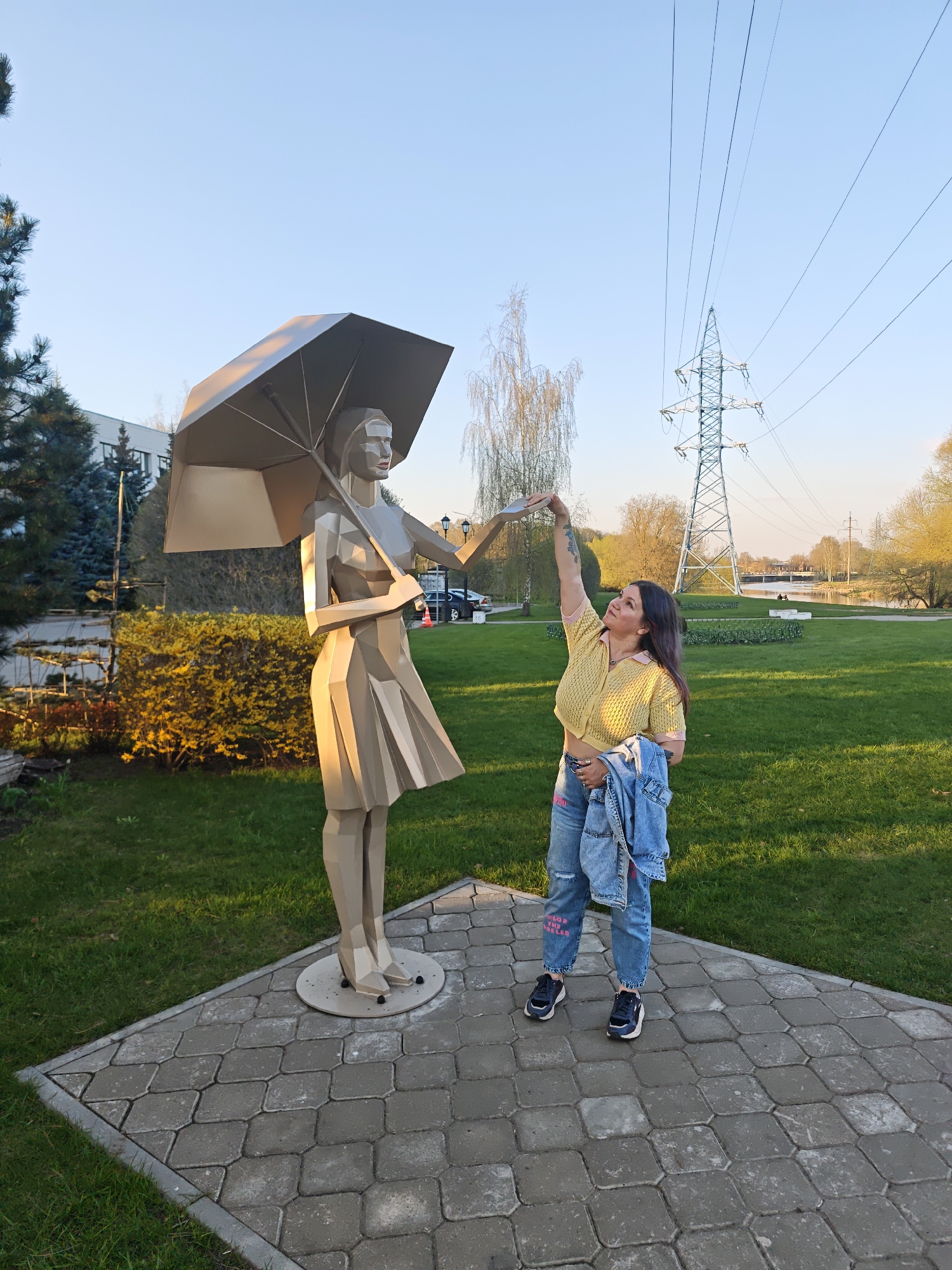 The image shows a woman interacting with a metal sculpture in a park. The sculpture depicts a woman holding an umbrella, rendered in a geometric style with flat planes. The woman in the image is standing next to the sculpture, with her right arm raised as if touching or interacting with the umbrella. She's wearing a yellow shirt, denim jeans with writing on them, and dark-colored sneakers. She also carries a denim jacket. They are both standing on a paved area. The background includes green grass, bushes, trees, a building, and an electrical power tower. Based on the available information, the location is identified as Noginsk, Russia.