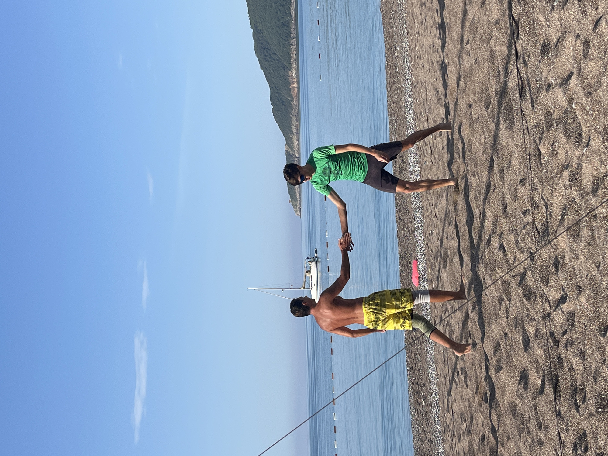 The image shows two men on the beach in Bečići, Montenegro, holding hands. The man in the green shirt and black shorts is facing the man in yellow shorts, who has a knee brace on his left leg. They are standing on a sandy beach with the Adriatic Sea and a distant mountainous shoreline in the background. A sailboat is visible on the water. A rope is stretched across the beach, appearing in the foreground. The sky is clear blue with a few thin clouds.