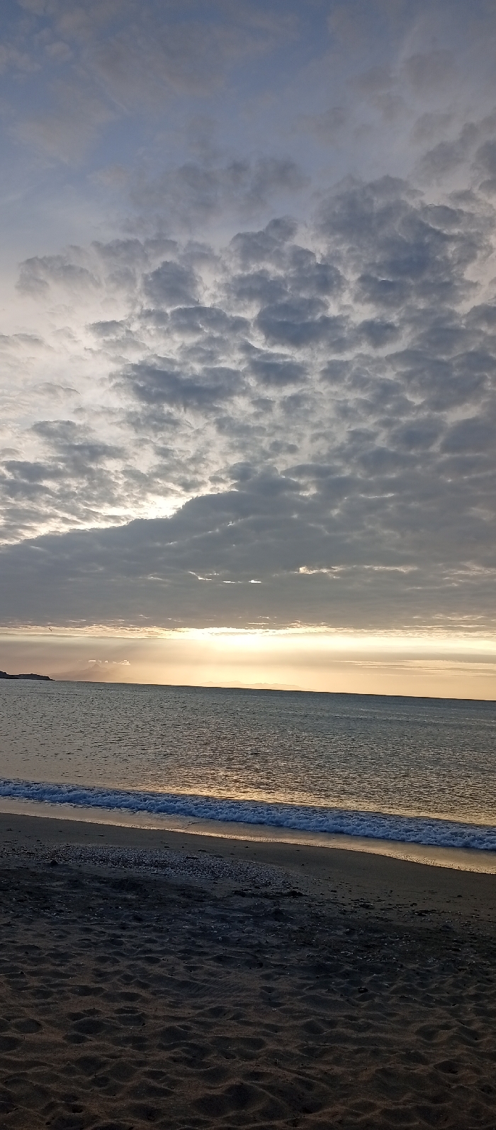 This image captures a coastal landscape in Juan Griego, Venezuela, during a low sun event. The upper two-thirds of the frame are dominated by an expansive sky filled with dense, grey stratocumulus clouds, with brighter, diffuse light breaking through patches near the horizon. A distinct band of intense, golden-yellow light extends horizontally across the middle of the frame, marking the horizon where the sky meets the ocean. The calm, dark grey-blue ocean surface reflects this golden light. In the mid-ground, small, white-crested waves gently break onto the sandy shore. The foreground consists of dark, damp sand, exhibiting subtle undulations and sparsely scattered with small, light-colored shell fragments or pebbles along the immediate waterline. A dark, low-lying landmass is visible on the far left, extending into the water. No people, animals, or artificial structures are discernibly present.