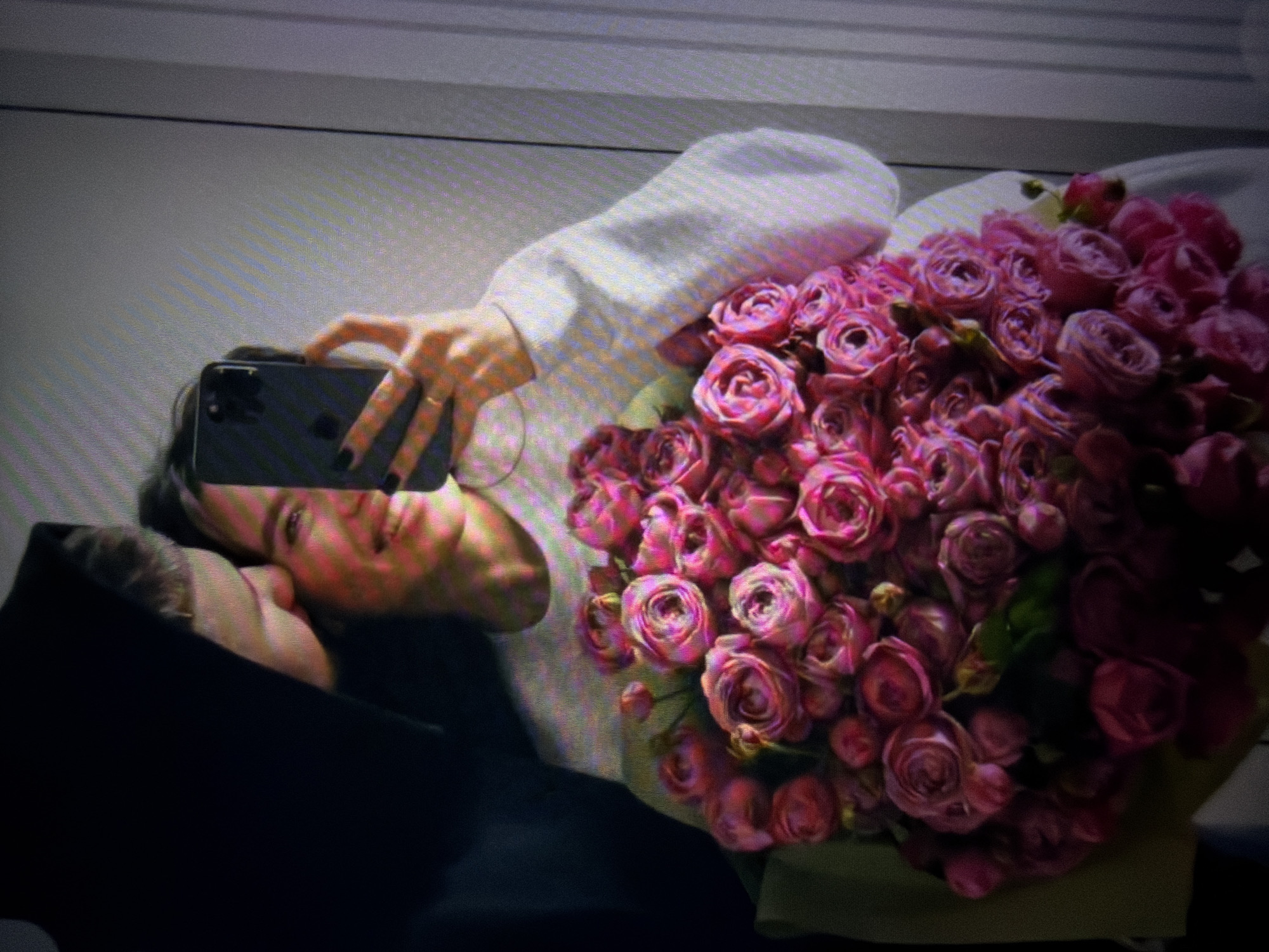 The media file captures a close-up, angled shot of two individuals and a large bouquet of pink roses. One person, with dark hair, is visible from the shoulders up, wearing a white top. They are holding a smartphone to take a photo, with their left hand angled towards the phone's screen and their right hand resting on the bouquet. A ring is visible on their left ring finger. The second person is mostly obscured by a dark hood or jacket, with only the side of their face and head visible, resting their cheek against the first person's shoulder. The overall mood appears intimate and affectionate.
The bouquet of roses is the dominant element in the right half of the frame, displaying numerous blooms in various stages of opening, with deep pink petals. The background is out of focus and appears to be an interior setting with a neutral-colored wall featuring horizontal lines, possibly blinds or a textured surface. The lighting suggests it could be daytime, but the indoor setting makes it difficult to determine the exact time of day or weather. The image is cropped from what seems to be a larger scene, possibly a mirror reflection given the angle and presence of the phone camera. There is no visible text in the media file. The provided location context of Beșalma, Moldova, places the scene geographically but does not offer specific visual cues within the image itself.