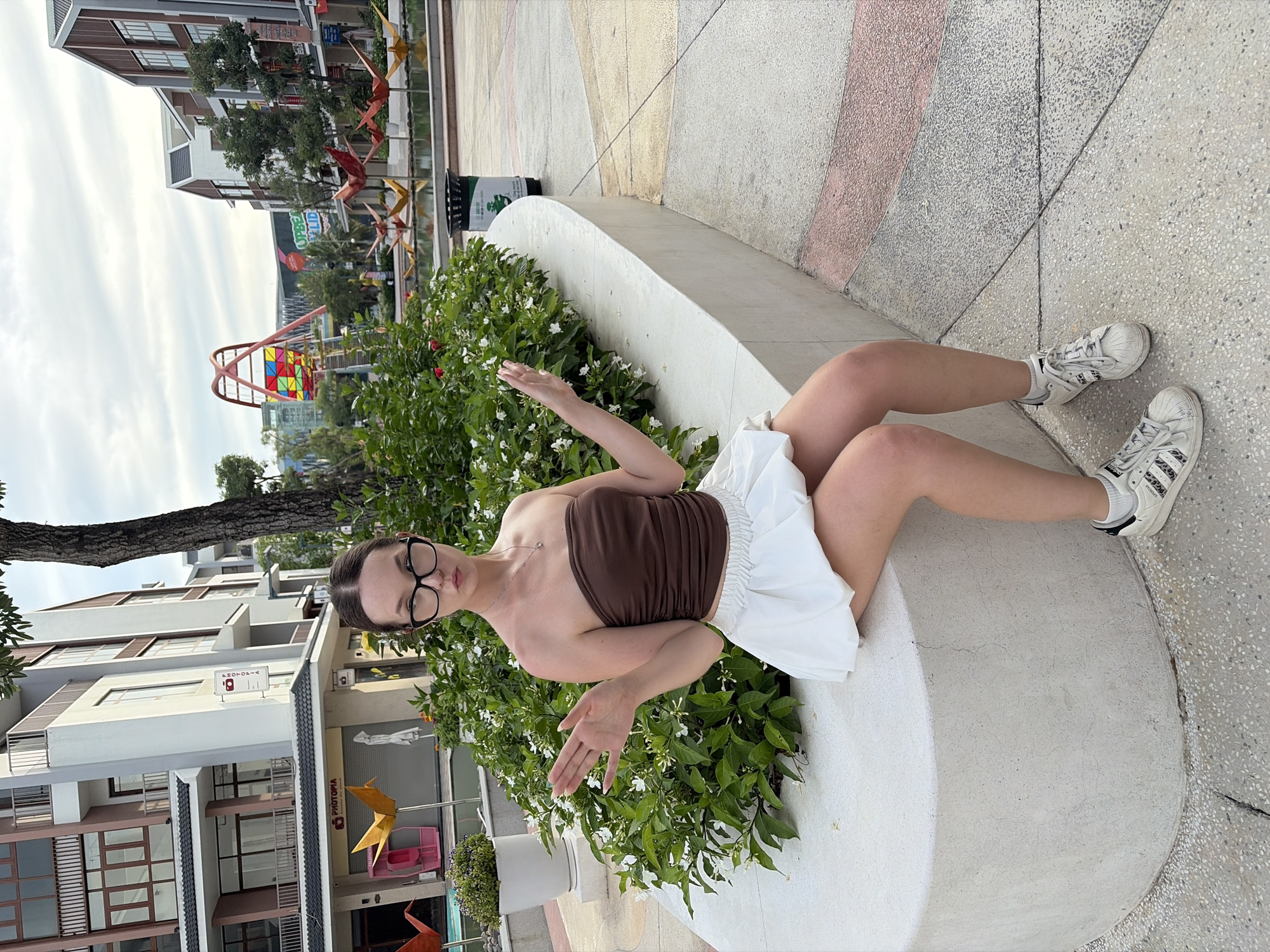 A young woman with dark hair pulled back and wearing glasses sits on a low, curved, concrete wall. She has on a brown strapless top and a white bubble skirt, and white sneakers with black stripes. Her arms are outstretched, palms up, in a questioning gesture. Behind her is a dense green bush with small white flowers and beyond that, buildings and an amusement park ride structure. The ground is paved with grey and pink tiles.