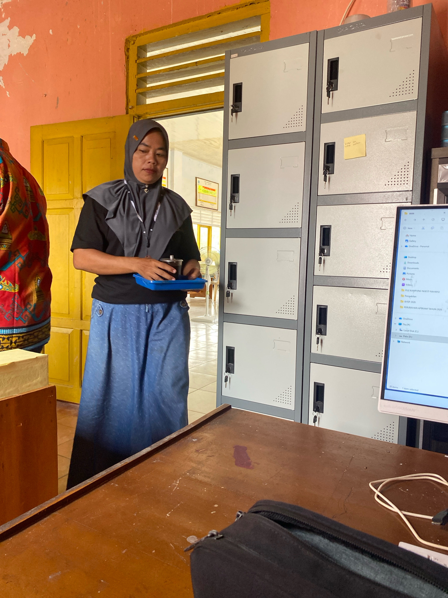 In an interior space with orange-painted walls and a yellow door, a woman wearing a gray hijab, black shirt, and blue patterned skirt holds a blue tray with a metal cup. To her right are several gray lockers with black handles and keyholes. A yellow sticky note is attached to one of the locker doors. In the foreground, a wooden desk is visible, with a black pouch and computer monitor on it. The scene appears to be in a classroom or office setting, possibly in the city of Mojo Pahit, Indonesia, given the attire and surrounding environment.