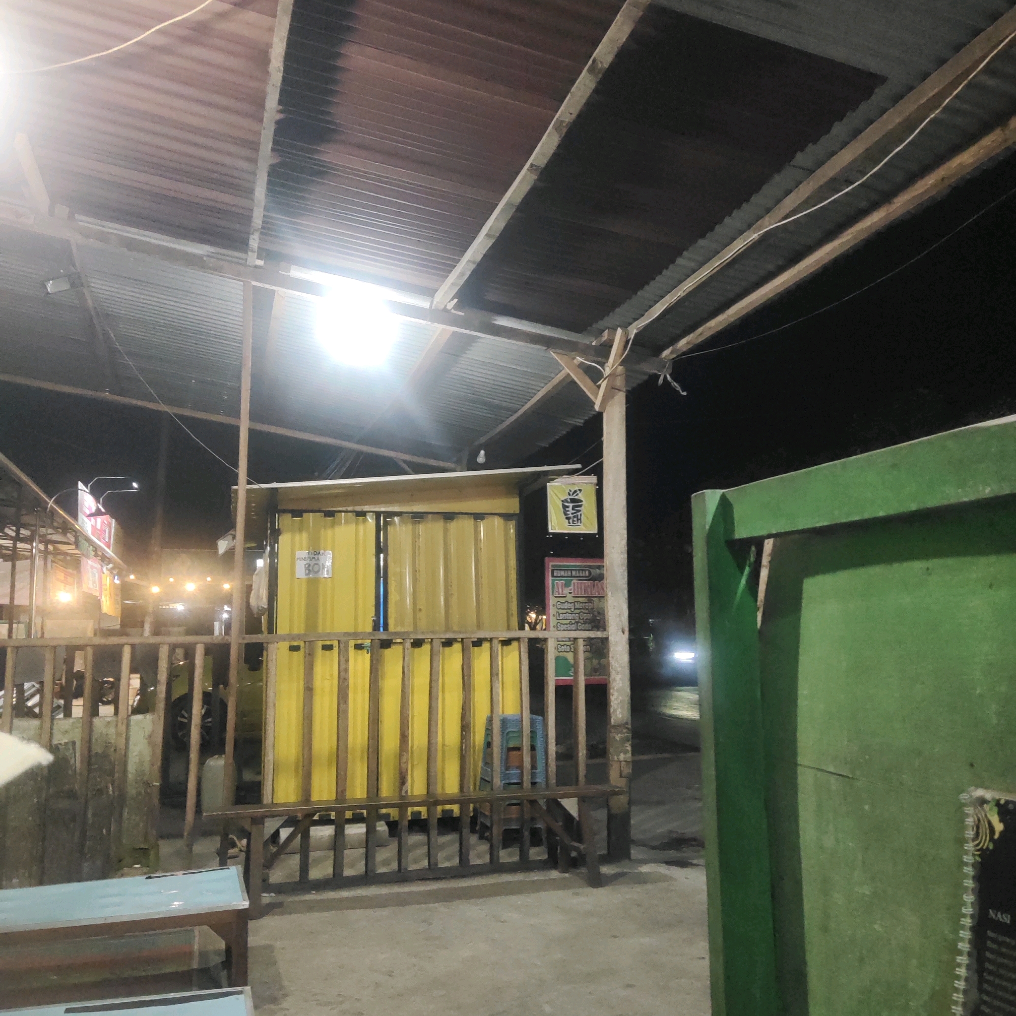 The image captures an outdoor, nighttime scene at what appears to be a roadside eatery or food stall in Sragen, Indonesia. The foreground features a wooden railing and two light blue benches. Behind the railing, a yellow corrugated metal structure, likely a food stall, is prominent. A small blue plastic chair is visible inside this structure. To the left of the yellow stall, other stalls or structures with colorful signage and lights are present, suggesting a marketplace or a row of food vendors. Further back, a vehicle with its headlights on is visible on a road. The overall environment is dimly lit, with overhead corrugated metal roofing supported by wooden and metal beams. The scene conveys a casual, potentially bustling, nighttime atmosphere common in such establishments in Indonesia.