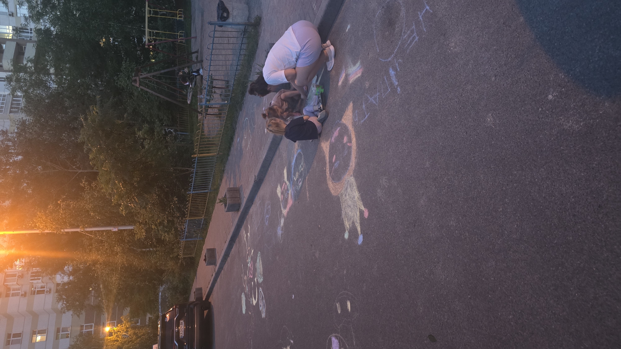 The image depicts a residential courtyard in Kyiv, Ukraine, during dusk. A group of people, including two young girls and an adult woman, are engaged in drawing with chalk on the asphalt. The chalk drawings are vibrant and varied, featuring stick figures, abstract shapes, and possibly words. The scene is set in front of a multi-story apartment building, with lush trees and a playground visible in the background. The warm glow of streetlights illuminates the area, casting long shadows on the ground. A black SUV is parked on the left side of the frame. The overall atmosphere suggests a peaceful, family-oriented evening activity.