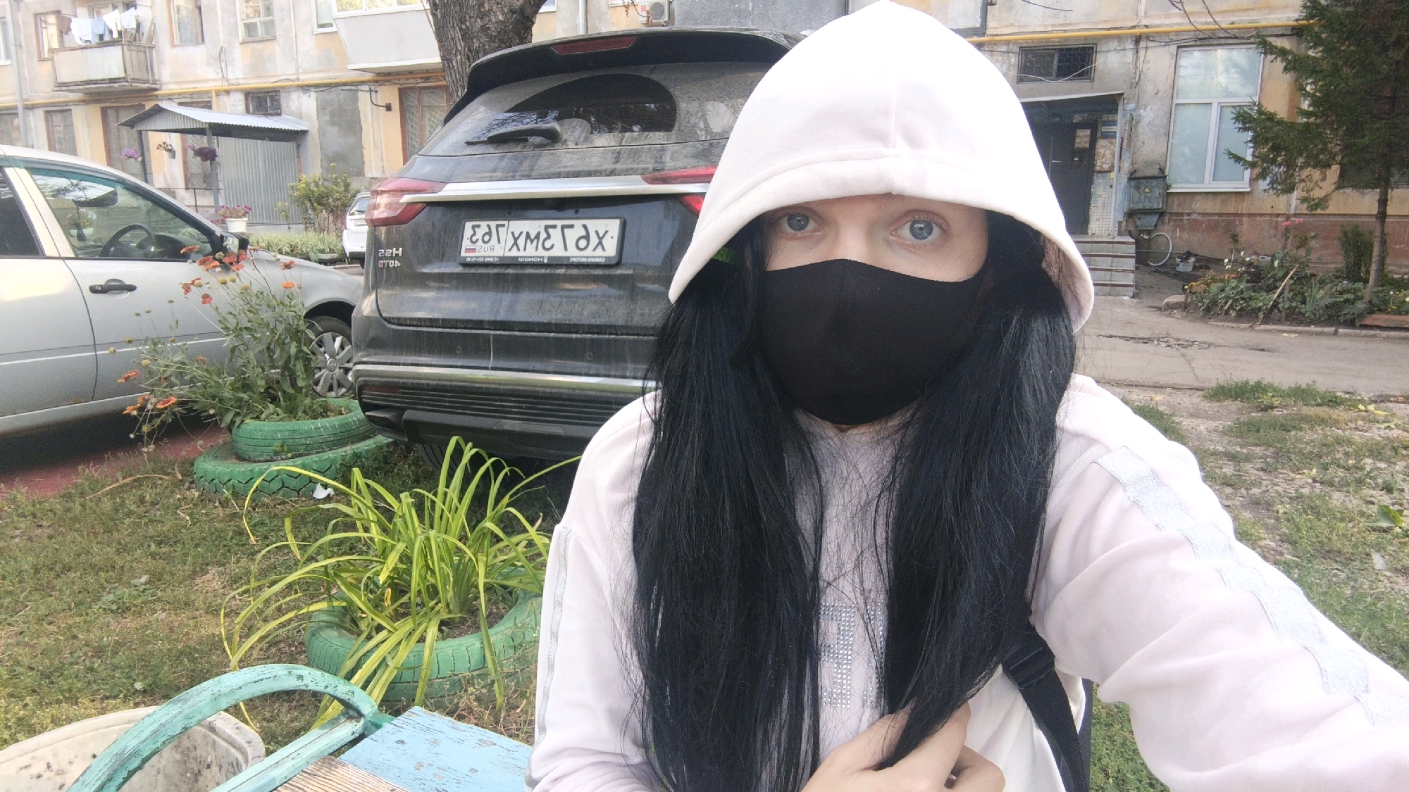 In Samara, Russia, a person wearing a light pink hooded sweatshirt, a black face mask, and a white hood stands in front of a dark-colored SUV. The SUV's license plate is visible and reads "EAT XMEГAX". To the left of the SUV is a silver sedan. The ground is covered in grass and dirt, with several green tires used as planters for orange flowers and green leafy plants. In the background, a multi-story apartment building with beige walls and balconies can be seen. The overall scene appears to be an urban residential area.