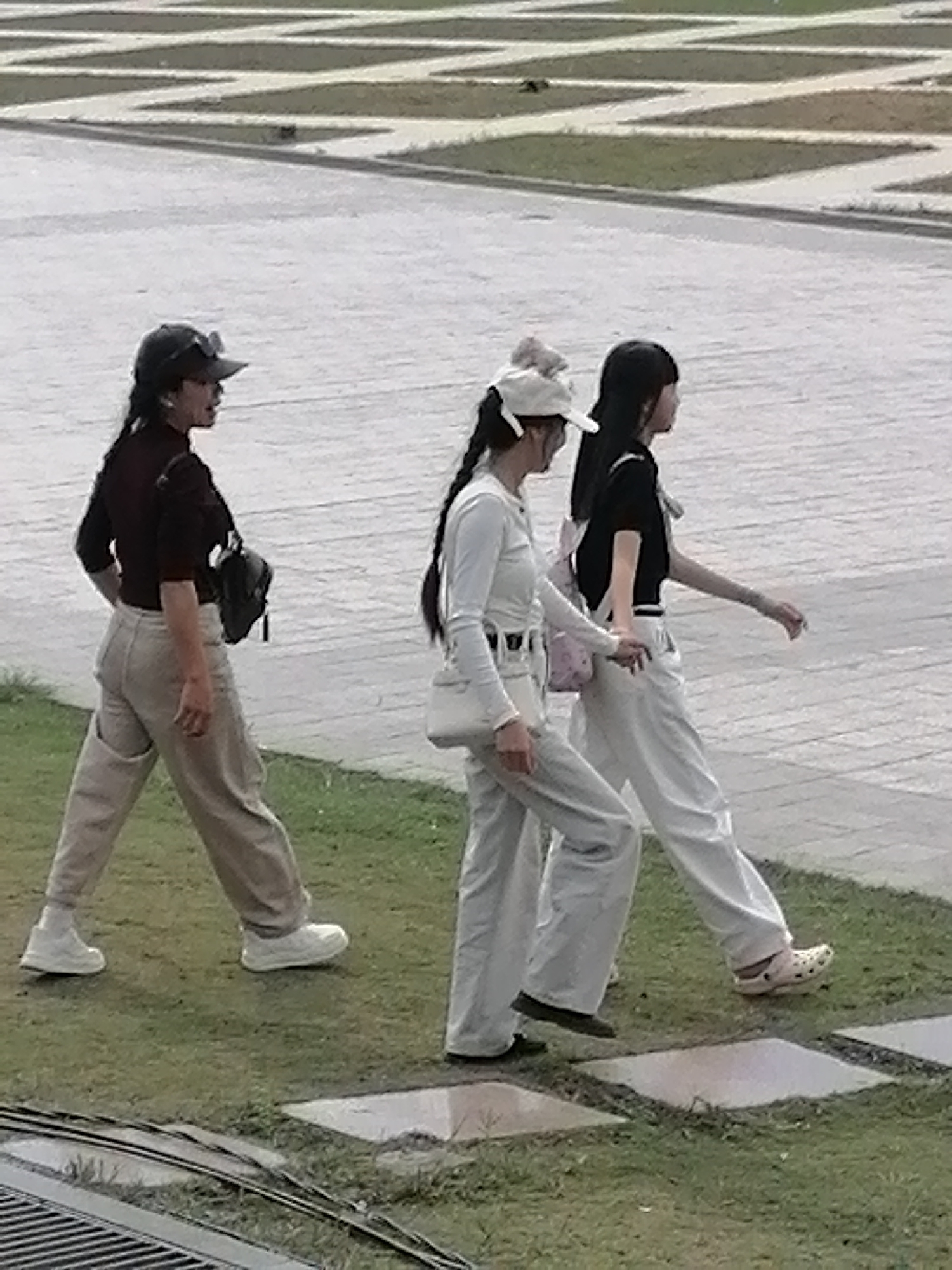 In Da Lat, Vietnam, three individuals are captured mid-stride on a paved area adjacent to a grassy patch. The foreground shows a metal grate structure. The first person, on the left, is wearing a dark maroon long-sleeved shirt, light-colored pants, and a dark baseball cap. A black bag is slung over their left shoulder. The second person, in the center, wears a white long-sleeved shirt, light-colored pants, and a white baseball cap. They are carrying a white crossbody bag. The third person, on the right, is dressed in a black short-sleeved top, white pants, and a light-colored backpack. The middle and right individuals are holding hands as they walk. The background features a patterned paved surface with intersecting lines and patches of green.