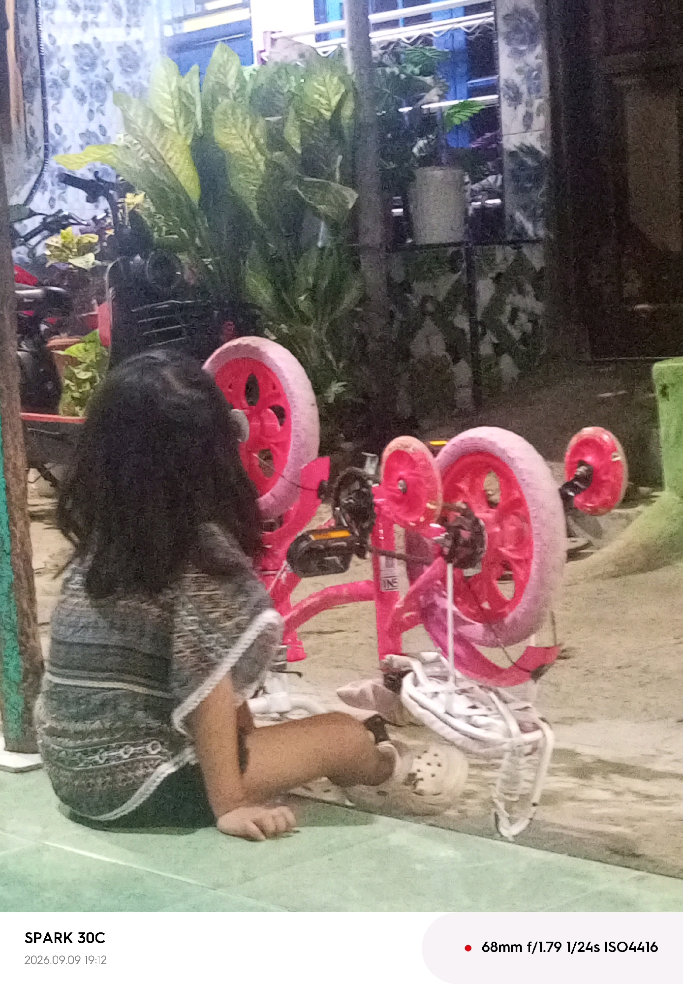 A young child with dark, shoulder-length hair is seated on a green surface, facing away from the camera. The child is wearing a gray and white striped shirt with a patterned hem. In front of the child is a pink tricycle with large wheels. The tricycle appears to be made of plastic and has a reflective strip on one of its wheels. White sneakers are attached to the pedals of the tricycle. The background is blurred but appears to include large green plants, a bicycle, and parts of a building. The lighting suggests it is either dusk or an indoor setting with artificial light. The text overlay indicates the image was captured by a SPARK 30C on 2026.09.09 at 19:12 with camera settings of 68mm f/1.79 1/24s ISO4416. The location is Tegal, Indonesia.