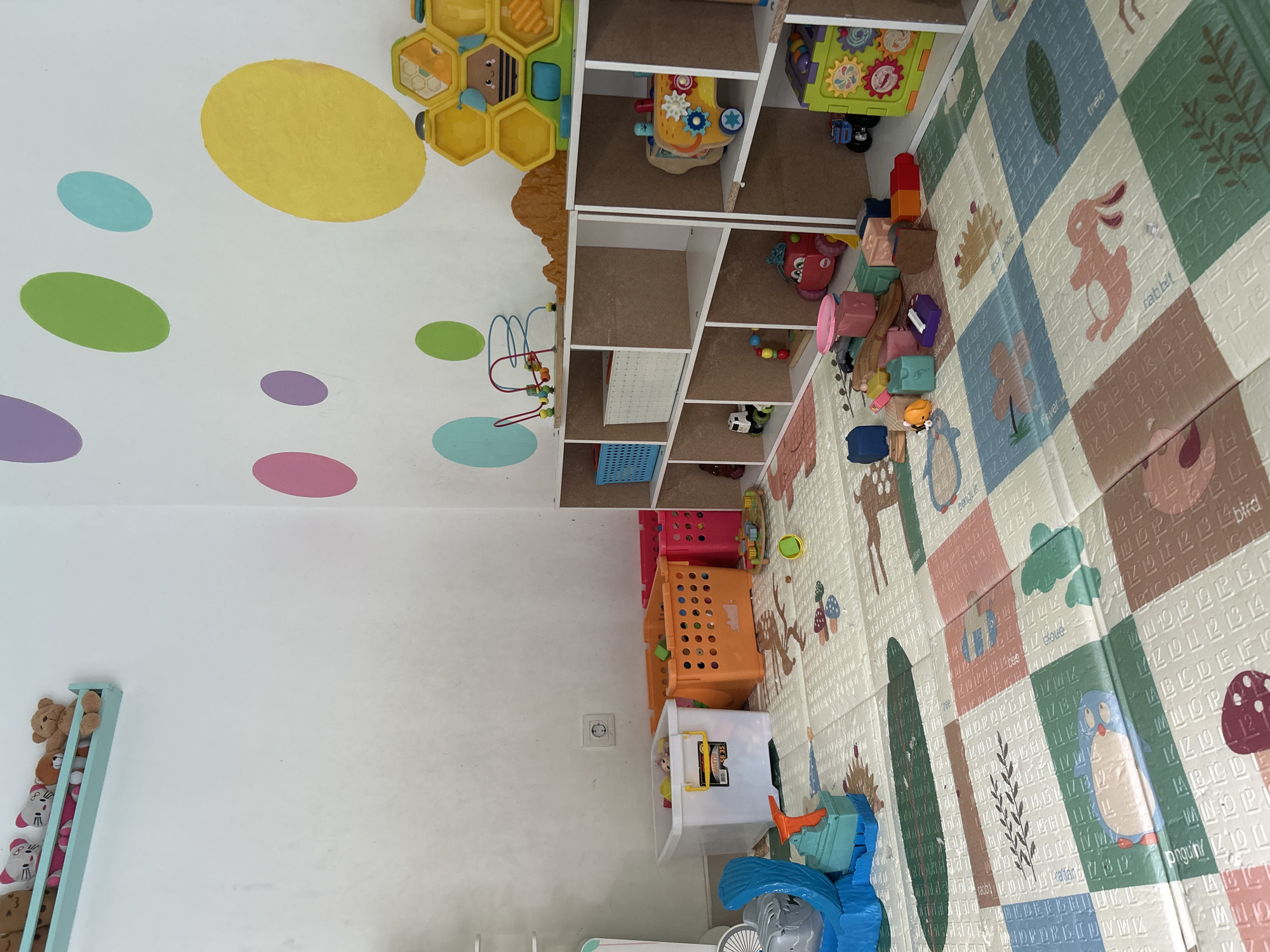 The image depicts a child's playroom in Kuta, Indonesia. The room is furnished with several shelves filled with toys, including stuffed animals, building blocks, and educational toys. A colorful play mat covers the floor, featuring alphabet letters and animal illustrations. Several storage bins are also visible, some filled with toys. The walls are decorated with large, brightly colored polka dots. The overall impression is one of a well-equipped and organized space for children's play and learning.