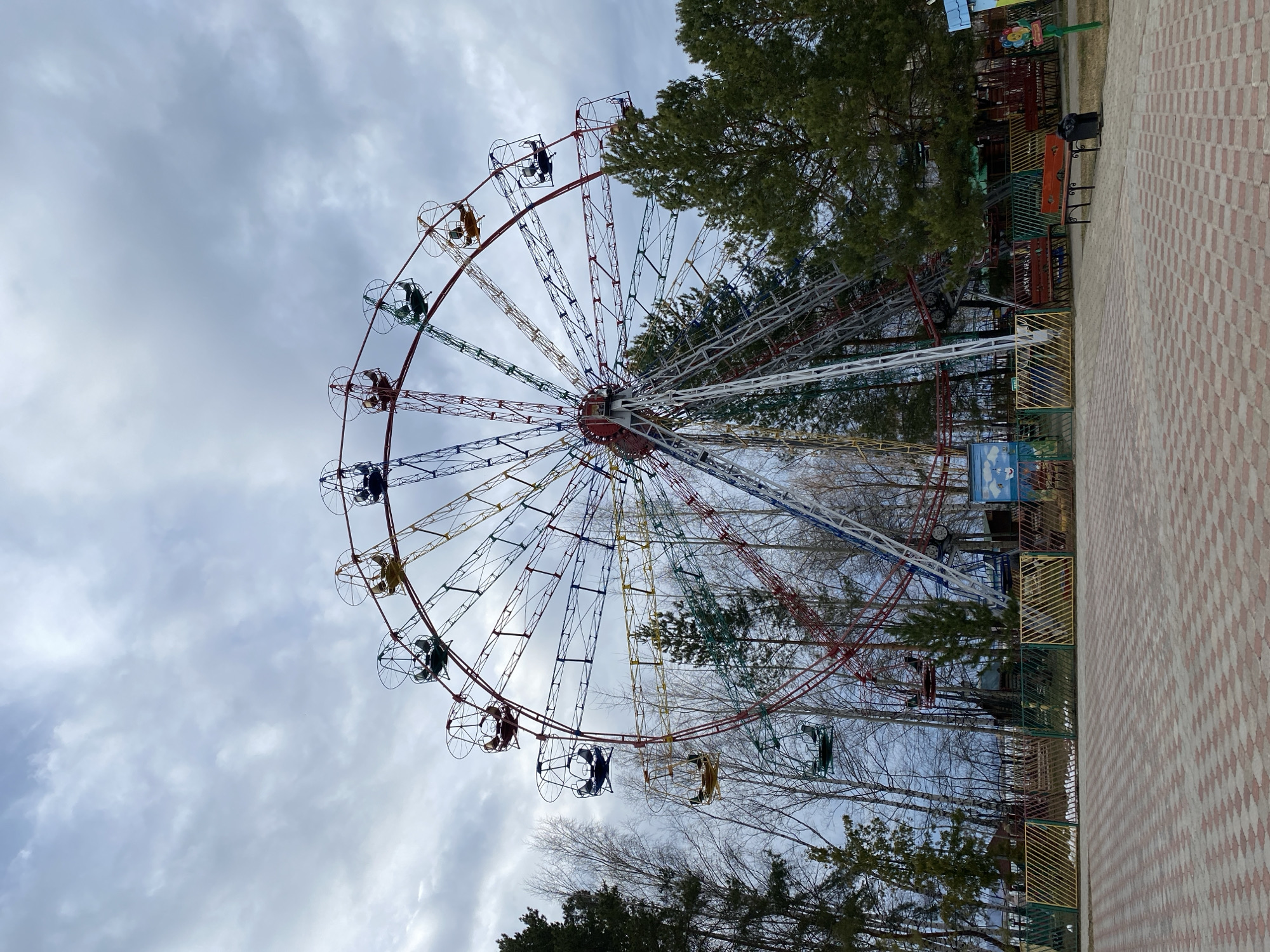A large, multi-colored Ferris wheel dominates the left side of the image, its intricate structure painted in segments of red, yellow, blue, green, and white. Numerous open-air gondolas hang from its perimeter, some appearing to contain dark shapes that might be people or objects. The Ferris wheel is set against an overcast sky filled with light gray clouds.
To the right, a wide paved path, constructed from alternating red and light-colored interlocking bricks, extends into the background. Adjacent to this path are several trees, including evergreen pines with dark green needles and bare deciduous trees, indicating a season like late winter or early spring. A wooden bench and a black trash can are positioned beside the path, near the trees. Sections of yellow and green fencing delineate areas around the Ferris wheel and along the path. In the background, glimpses of other amusement park structures or signs are visible through the trees, though any text on them is illegible.
The scene depicts a quiet amusement or public park, likely located in Chaikovsky, Russia. The absence of direct sunlight and the bare trees contribute to a subdued atmosphere. No clear activity is taking place. Visible text on signs is too indistinct to be read.