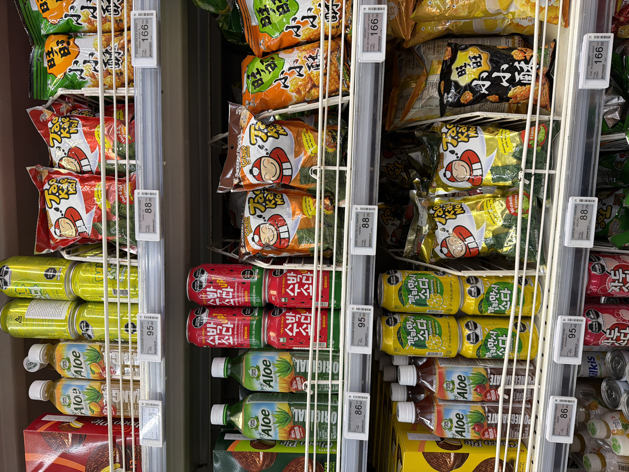 The image displays a shelf in a supermarket located in Sokilnyky, Ukraine, stocked with various food and beverage items. The shelves are filled with an assortment of products including bottled aloe vera drinks in green and brown hues, canned sparkling beverages in red and yellow packaging featuring Korean script, and bagged snacks. Some of the snack bags have red and white cartoon characters on them, while others have orange and yellow packaging with Chinese characters. Price tags are visible on some of the items. The lighting suggests an indoor commercial environment. There are no people visible in the frame.