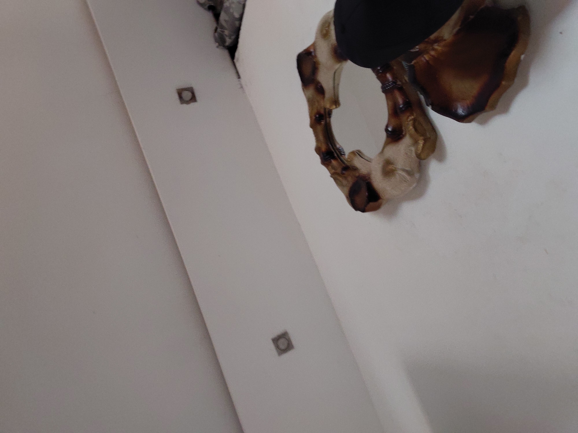 The image displays an indoor scene, primarily featuring a white wall or ceiling surface with a distinct diagonal line, possibly indicating a corner or a seam between two structural elements. Mounted on the upper right side of this surface is a decorative object, which appears to be a mirror or an ornamental frame. This object exhibits a rustic aesthetic, characterized by irregular, organic shapes and textures, with colors ranging from deep brown to light tan, suggestive of aged wood, antlers, or a naturally formed material. A section of a solid black object is visible at the very top edge, partially obscuring the decorative item.
Further to the left, embedded within the white surface, are two small, square-shaped fixtures. One is located in the upper-left quadrant, and the other is positioned closer to the center-left. These appear to be recessed light fixtures or electrical outlets. The lighting is uniform, suggesting an interior space, and there are no discernible shadows that would indicate a specific time of day or external natural light source. No human figures or activities are depicted. No text is visible in the image. The scene is static and devoid of overt emotional cues. The location context for this indoor scene is Santa Ana, Venezuela.