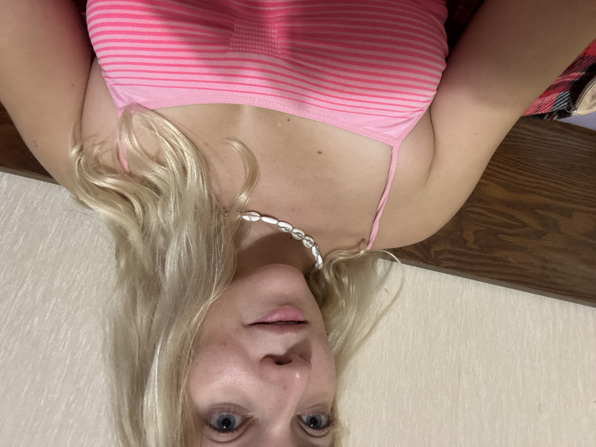 A blonde woman with blue eyes is pictured in a close-up selfie. She is wearing a pink and white striped tank top and a necklace made of shell-like beads. The background appears to be a textured wall and a wooden surface, possibly a headboard. The lighting suggests an indoor setting. The image provides no information about the city of Minsk or Belarus.