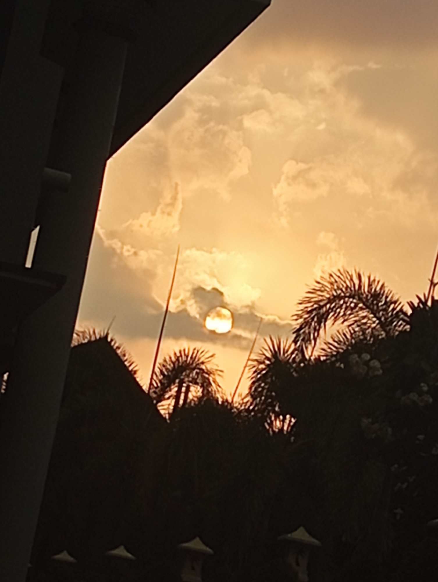 The image shows a sunset over a tropical environment, likely in the city of Mataram, Indonesia. The sun is partially obscured by clouds, casting a warm, orange glow across the sky. In the foreground, a dark silhouette of a modern building's overhang is visible on the left, with a portion of a pillar extending downwards. To the right, the dark silhouettes of palm trees and other lush vegetation create a dense canopy. The setting sun, a bright, glowing orb, is visible through a gap in the clouds. The overall impression is one of a serene and tranquil evening in a tropical locale.
