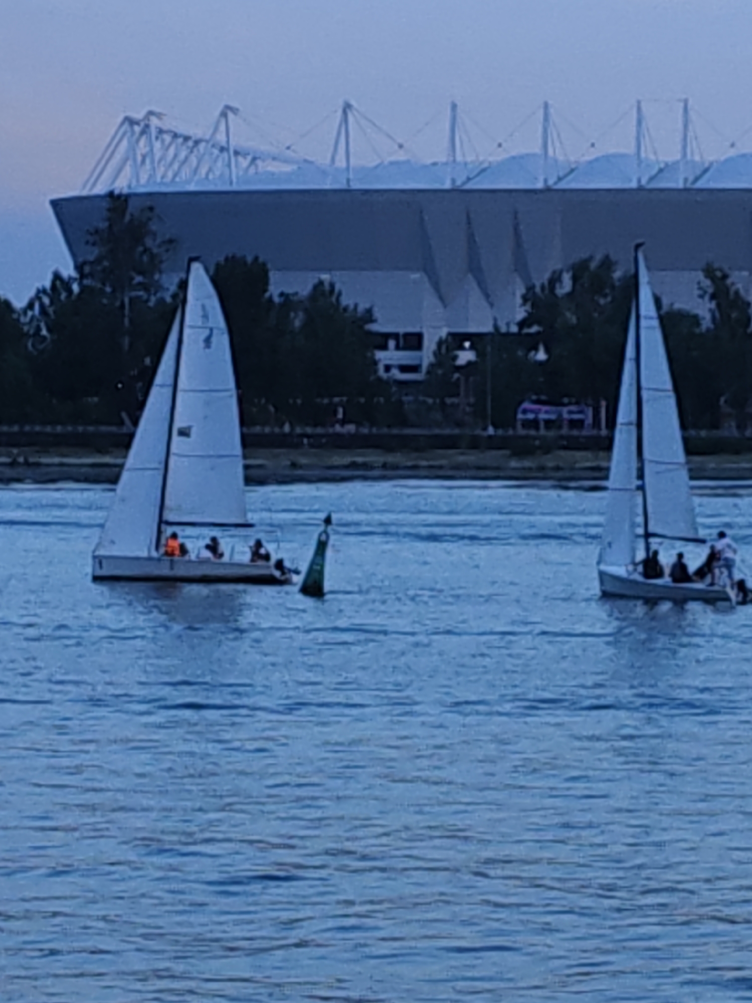 In the foreground, two sailboats navigate a calm body of water. The sailboat on the left has at least four people on board, with one person wearing an orange life vest. A green buoy with a white top floats near the left sailboat. The sailboat on the right also has several people visible. In the background, a large, modern stadium with a distinctive white, angular roof structure is prominent. Trees line the shore between the water and the stadium. The sky is a pale, hazy blue, suggesting it may be early morning or late evening. The overall scene depicts a serene water activity with a large architectural landmark in the distance.