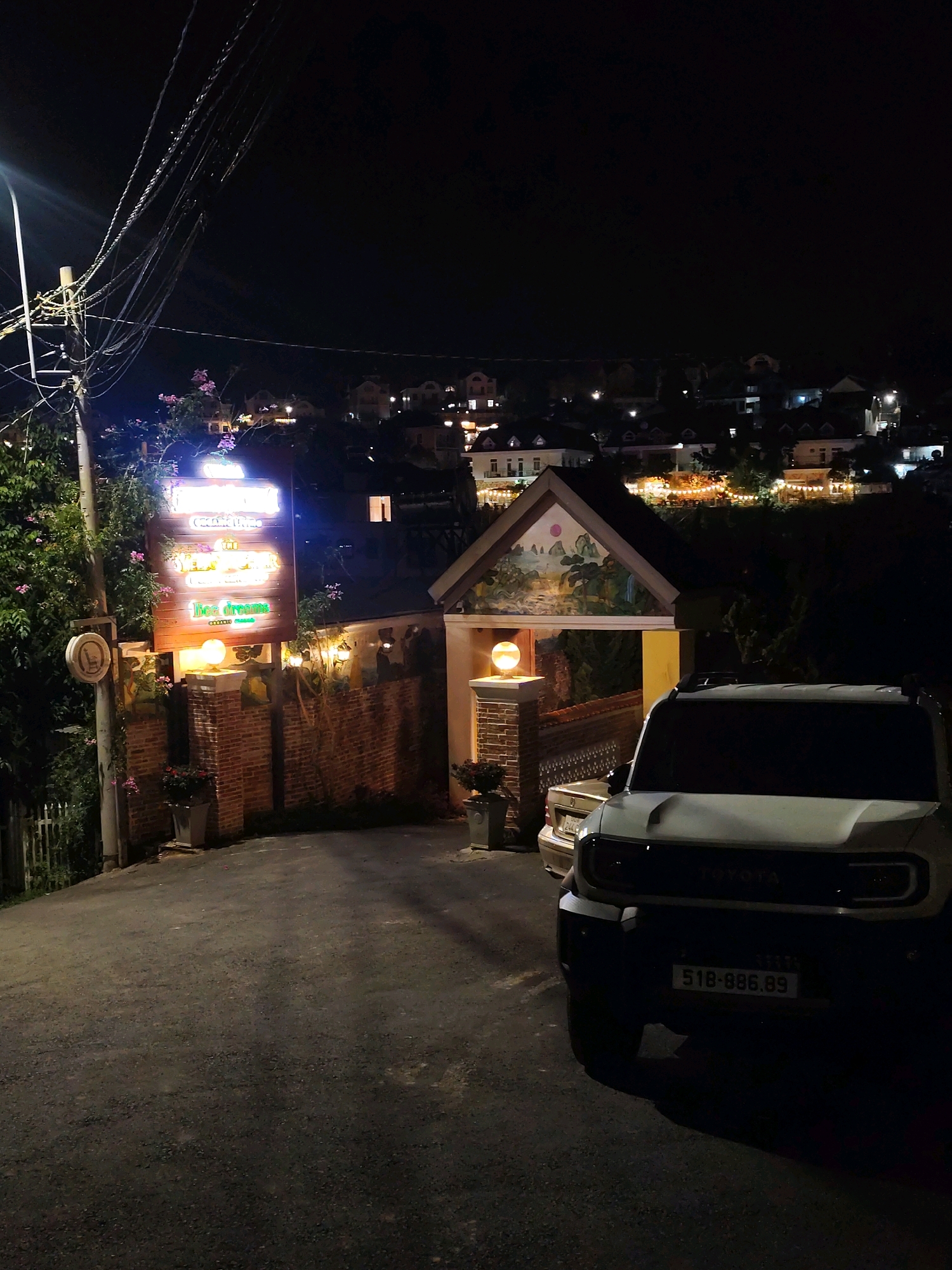 At night, a paved road curves uphill towards a gated entrance of what appears to be a guesthouse or restaurant. The entrance features a brick wall on the left, a gated archway adorned with a mural of a landscape, and a warmly lit spherical lamp on a pedestal. Above the brick wall, a wooden sign with neon lettering reads "Bee dreams" in green, and above that, "The Garden" in pink, and then "Organic Villa" in white. A white SUV is parked in the foreground, with its rear license plate visible, reading "518-886.89". In the background, the lights of the city of Da Lat, Vietnam, twinkle across the hilly landscape, with a few buildings visible on the slopes. The sky is dark, and overhead, tangled electrical wires are strung between utility poles.