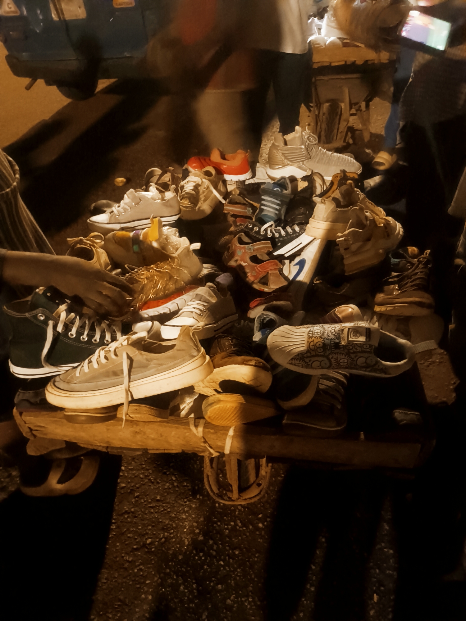 A street scene in Bukuru, Nigeria, at night depicts a makeshift shoe stall. A large pile of various types of shoes, primarily sneakers and casual shoes, are arranged on a wooden platform supported by wheels. A person's hand is reaching towards the shoes. Other people are standing around the stall, with one person holding a mobile phone. A vehicle is partially visible in the background. The lighting is dim, creating shadows across the scene.