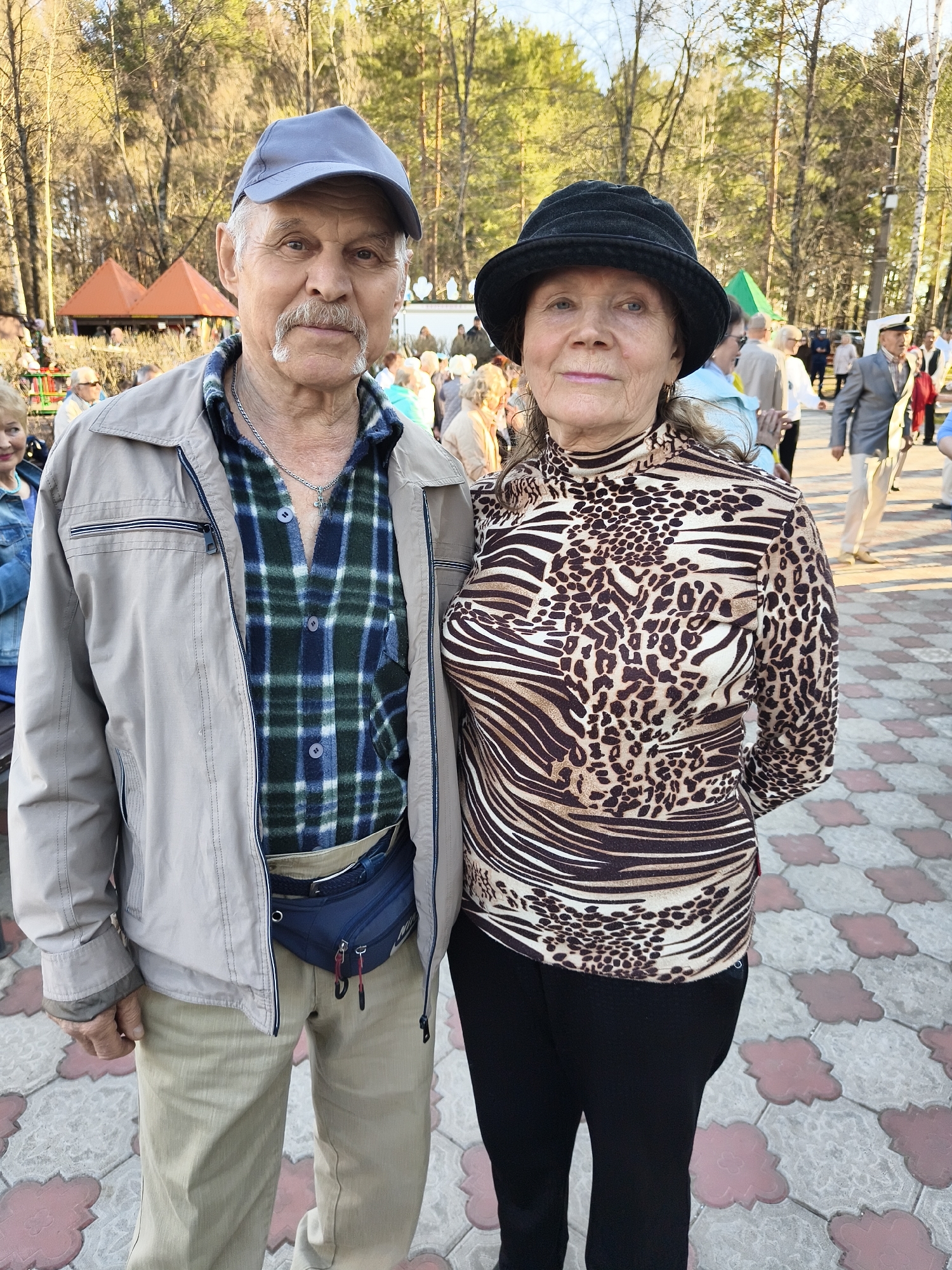 In a public area within the city of Unknown, Unknown, a man and a woman stand close together, facing the camera. The man is wearing a light grey jacket over a green and blue plaid shirt, with a blue cap on his head. A blue waist pack is strapped around his waist. The woman, to his right, is dressed in a brown and white animal print turtleneck shirt, paired with a black hat. She wears black pants. The background contains a crowd of people, trees, and tents. The ground is paved with patterned tiles.