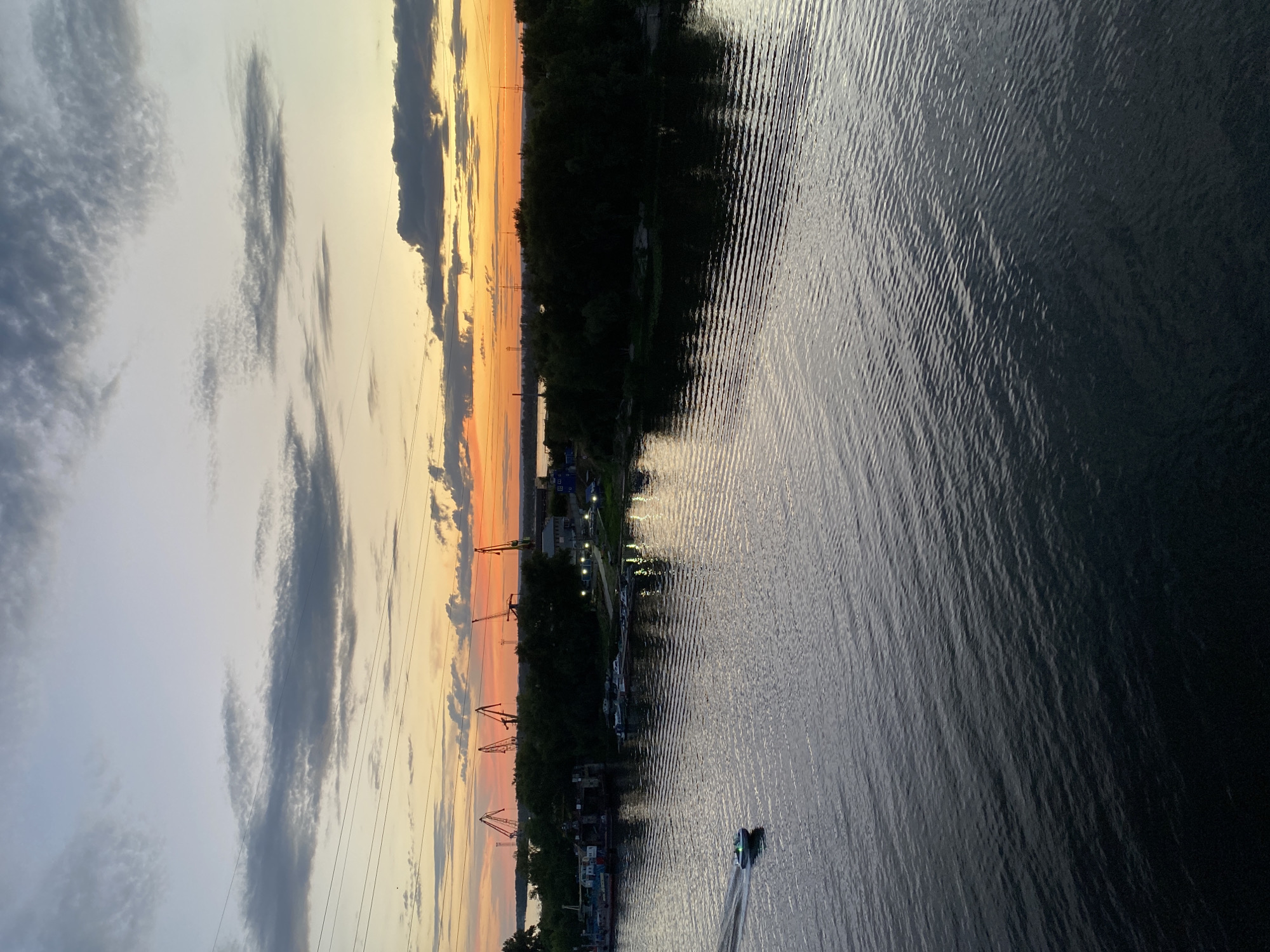 This is a landscape photograph taken from a high vantage point, looking down on a body of water and the shore. The image is oriented vertically. The sun is setting, casting a warm orange and yellow glow across the sky, with scattered clouds in shades of grey and white.
On the left side of the frame, the sky dominates, transitioning from a pale blue at the top to the vibrant sunset colors near the horizon. Thin, dark power lines stretch horizontally across the sky, indicating the presence of infrastructure.
The right side of the image is covered by a dark, rippling body of water, likely a river or lake. The water's surface reflects the sky and the dark silhouette of the shore. A small boat is visible in the water, leaving a white wake behind it as it moves.
The shore is lined with dark green trees, creating a dense border between the sky and the water. Along the shoreline, there are several structures, including what appear to be cranes, docks, and buildings, suggesting a port or industrial area. Some of these structures are illuminated by lights. The presence of multiple boats docked along the shore further supports this interpretation.
The overall mood of the image is serene and tranquil, characteristic of a sunset over water. The time of day is clearly evening, and the weather appears clear with some cloud cover. The location context provided suggests this scene is in Novokuybyshevsk, Russia. No visible text is present in the image.