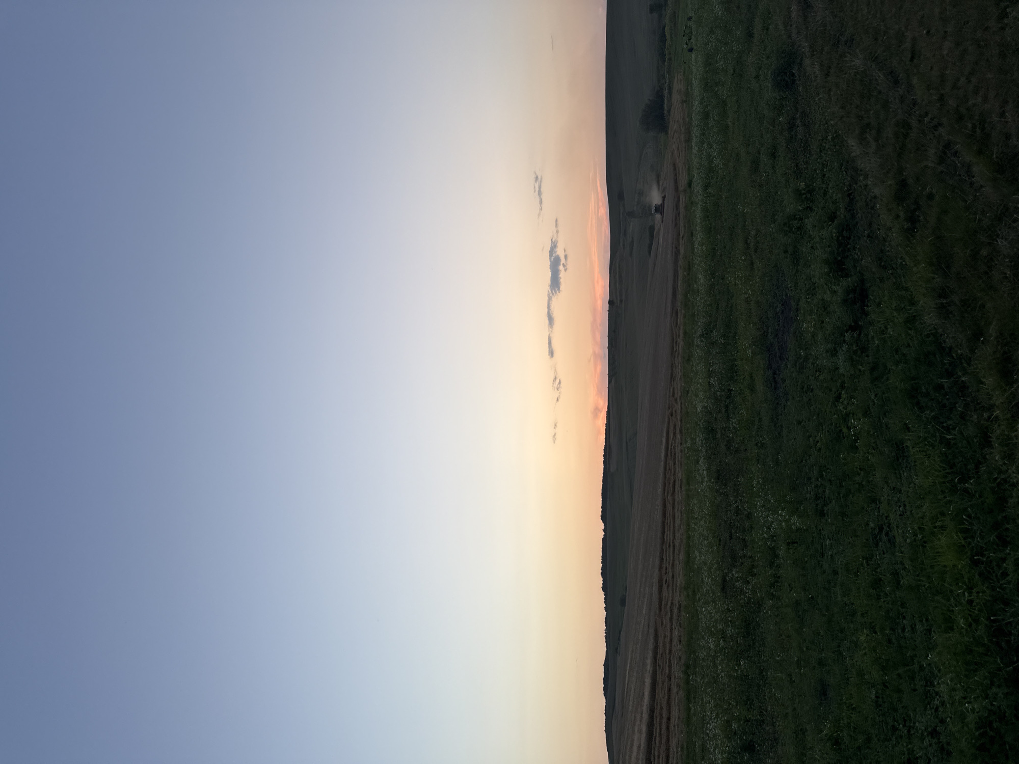 A wide-angle vertical shot captures a rural landscape at dusk. The sky dominates the upper portion of the frame, transitioning from a pale blue at the top to a warm orange and pink near the horizon. A few wispy clouds are scattered across the sky.
Below the sky, rolling hills form the backdrop. The closest hill is covered in dark green grass, with some lighter patches of wildflowers. Further in the distance, a harvested field is visible, appearing as rows of dried straw. A combine harvester, a yellow and green machine, is operating in this field, kicking up a trail of dust. Another piece of agricultural equipment is parked on the right side of the frame. The scene is quiet and serene, suggesting the end of a day's work in the countryside.