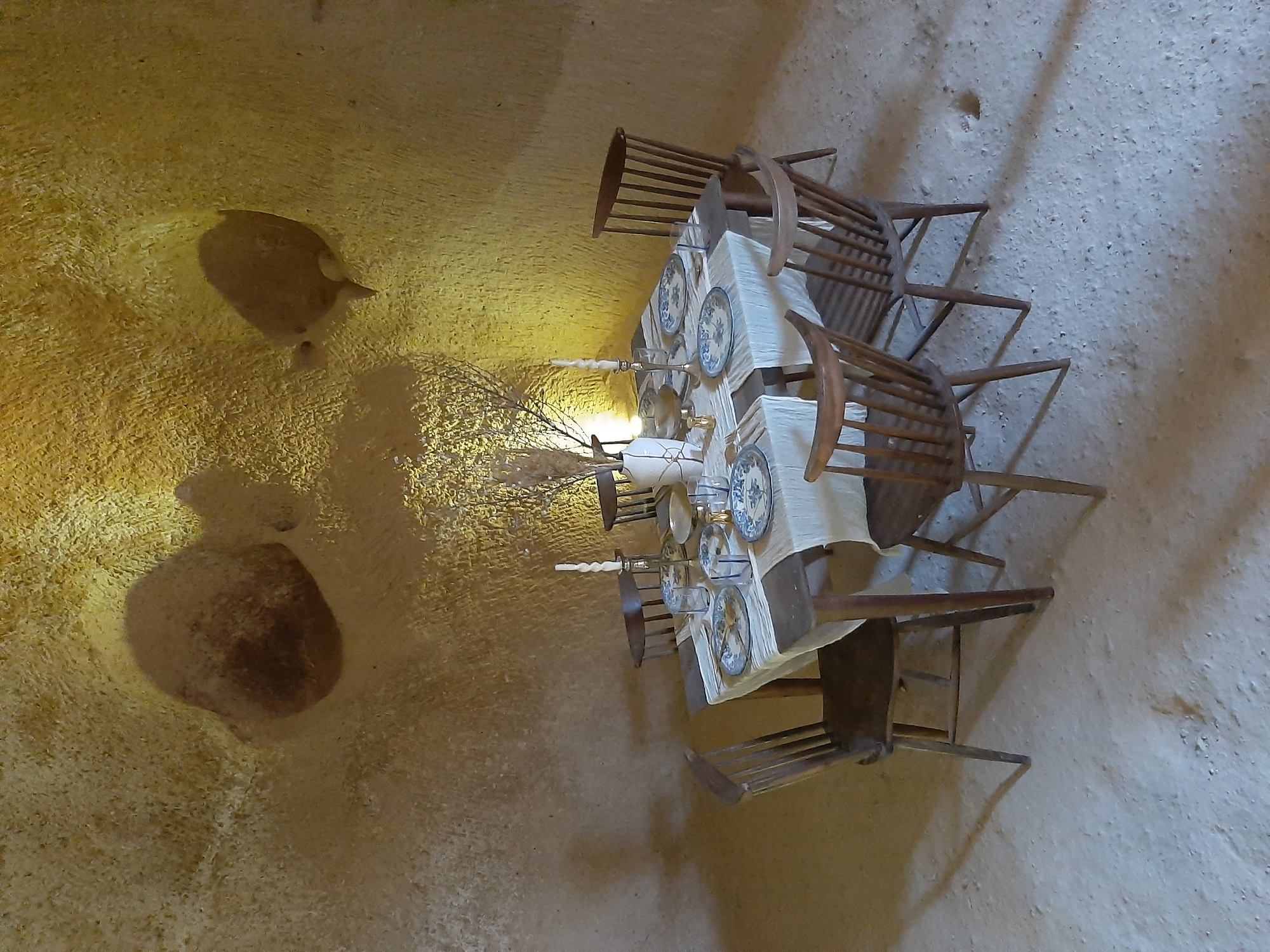 The image depicts an indoor setting with a table and chairs, possibly in a cave-like environment given the rough, textured walls and ceiling that appear to be made of natural rock. The lighting is warm and yellow, casting shadows on the walls and floor.
In the center of the scene is a rectangular table covered with a white tablecloth. The table is set for a meal with several plates, cutlery, glasses, and a centerpiece. The plates have a blue and white floral pattern. A tall, thin white candle in a candlestick is visible on the table, along with a vase containing dried foliage.
Six wooden chairs surround the table. The chairs have a spindle back design and appear to be made of dark wood. The floor is covered in a light-colored, granular material, resembling sand or fine gravel.
The overall impression is a rustic yet elegantly set dining area within an unusual, natural architectural space. There are no people visible in the image. The environment suggests a historical or unique lodging or dining establishment.