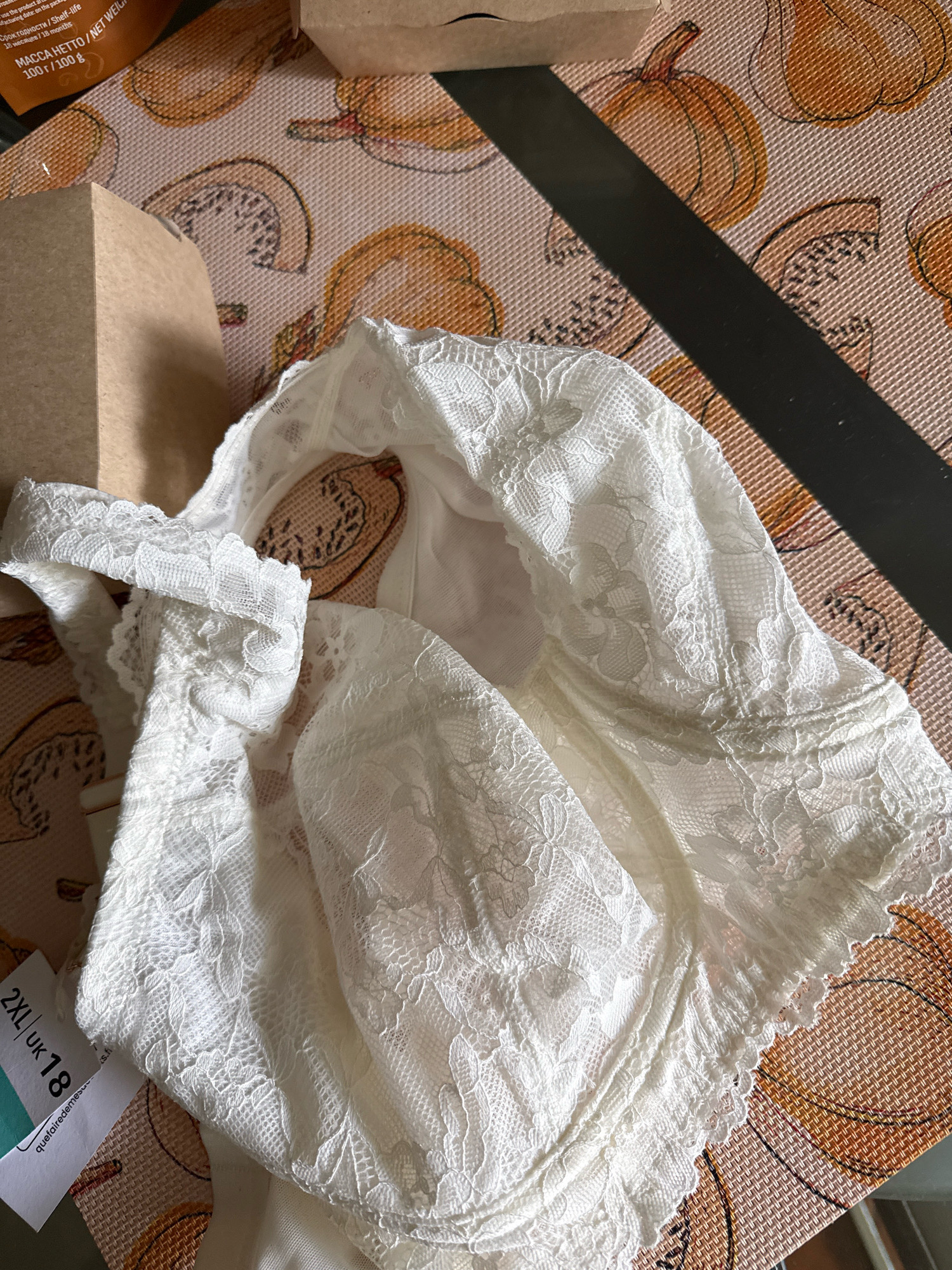 The image shows a white lace bra laid out on a placemat with a pumpkin pattern. The bra is partially folded, with a size tag visible on the left side that reads "2XL UK 18". A cardboard box is positioned to the left of the bra. The background consists of a dark surface with a striped pattern. The lighting suggests an indoor environment. The scene is devoid of people and any specific location in Kaliningrad, Russia cannot be determined from the visual information provided.