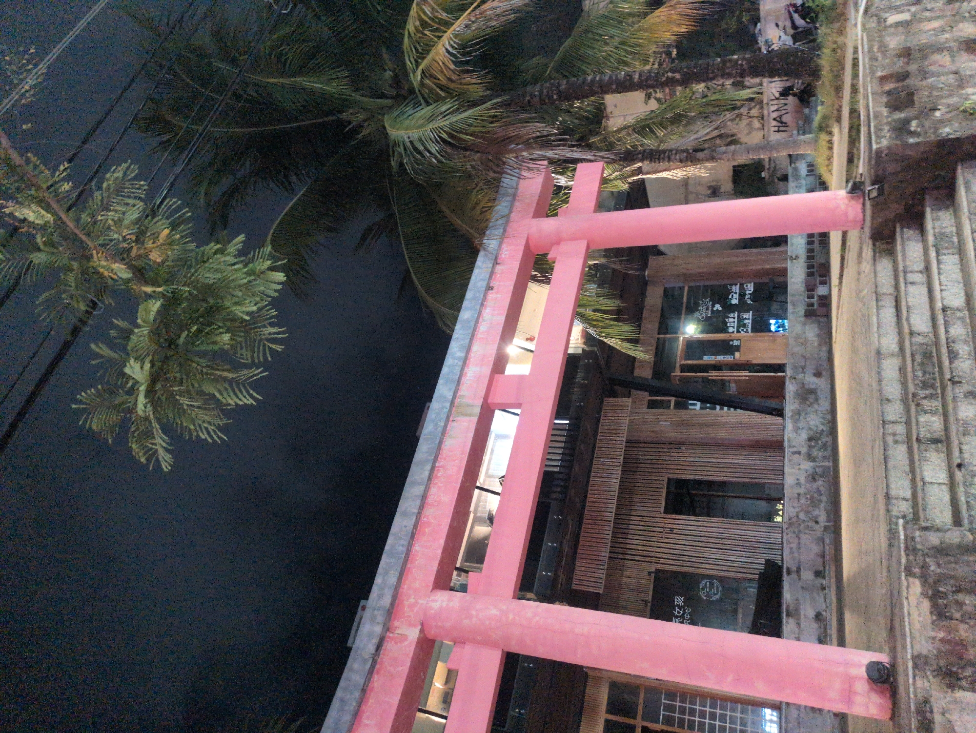 A building behind a pink torii in Phu Quoc, Vietnam. The pink torii is constructed of square beams connected with rounded joints. A palm tree is visible behind the torii, and the sky is dark, suggesting it is night. The building behind the torii has a wooden facade with windows and a sign in Vietnamese. The building is to the right of the torii, and the torii is in the foreground. A set of stairs runs along the right side of the image. The ground is dark and the image is slightly blurry.