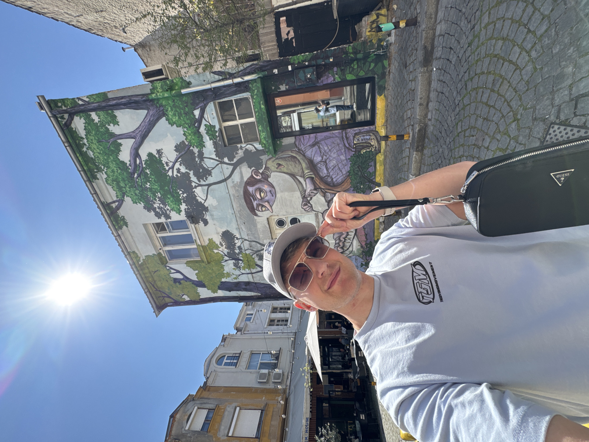 A man wearing a grey cap, sunglasses, and a light-colored t-shirt with black lettering stands at an angle to the camera, holding a black bag on his shoulder. Behind him is a building with a large mural depicting a tree and a whimsical figure. The buildings in the background suggest a city street with shops and restaurants. The cobblestone pavement and architectural style suggest that the location is in Plovdiv, Bulgaria, specifically in the artistic district of Kapana.