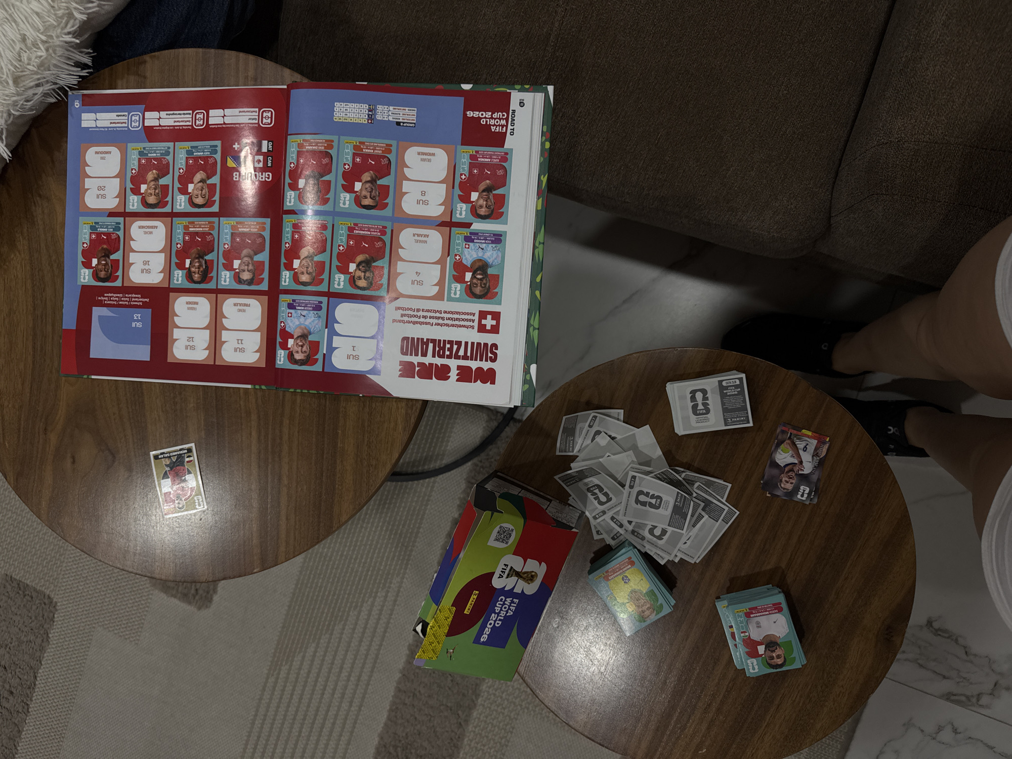The image shows an open sticker album for the FIFA World Cup 2026. The album is open to the Switzerland section. Various FIFA World Cup 2026 sticker packs and loose stickers are scattered on a round table alongside the album. A 'Mohamed Salah' sticker is on a separate round table. A person's legs are visible near one of the tables. The setting appears to be a living room or similar indoor space. The location is not provided.