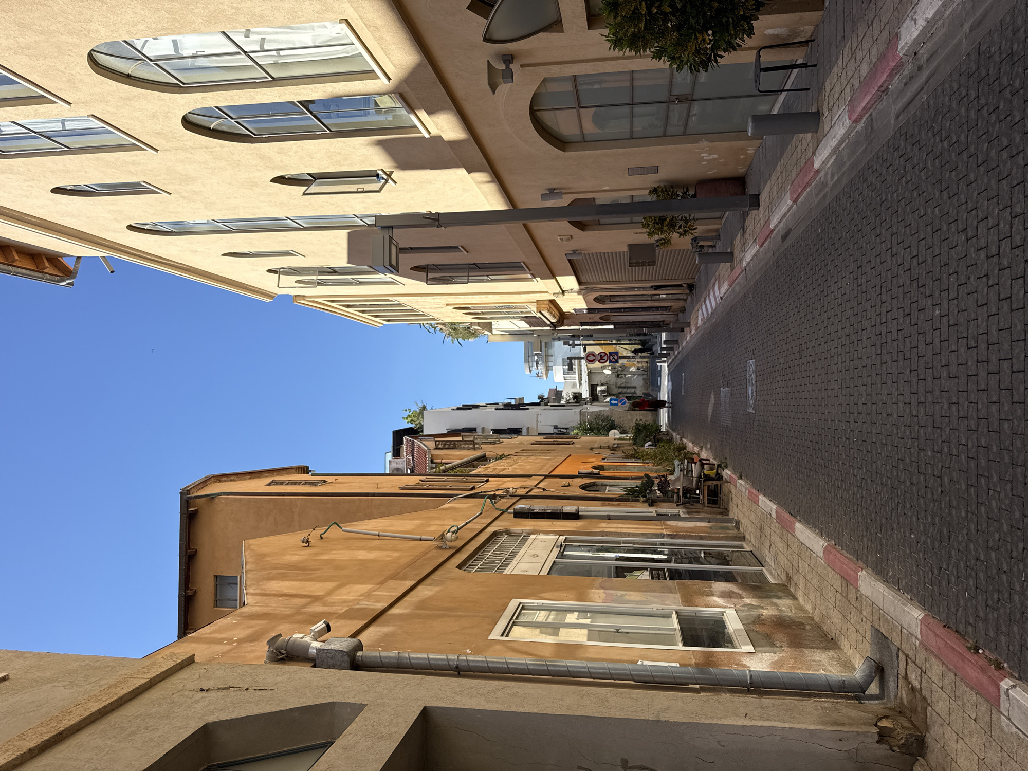 A narrow, cobblestone street in Tel-Aviv, Israel, is flanked by tall, ochre-colored buildings with arched windows. The street is well-lit by sunlight, casting shadows on the ground. A single person, wearing a red top and dark pants, walks away from the camera in the distance. The buildings on the left have a more weathered appearance, with visible pipes and window grates, while the buildings on the right are smoother and more modern. The street is lined with a sidewalk that has a red and white striped curb. The sky is clear and blue.