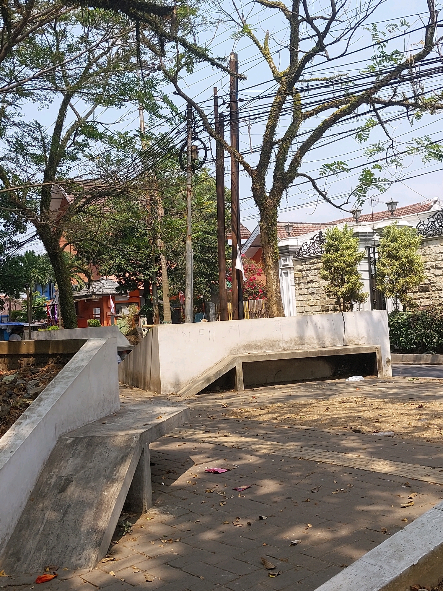 The image depicts a street scene in Malang, Indonesia, featuring a concrete bench and walkway on the right side, with a tree-lined street and buildings in the background. The walkway is paved with dark gray bricks and scattered with fallen leaves. The concrete bench is a long, rectangular structure with a hollowed-out section underneath, appearing to be a built-in seating area. In the background, a building with a white stone facade and decorative ironwork is visible, along with a red-orange building with a tiled roof. Lush green trees with moss-covered branches dominate the upper portion of the image, with numerous electrical wires crisscrossing between them. The sky is a clear blue. There are no people clearly visible in the foreground. The overall impression is one of an urban outdoor space with a mix of natural elements and man-made structures.