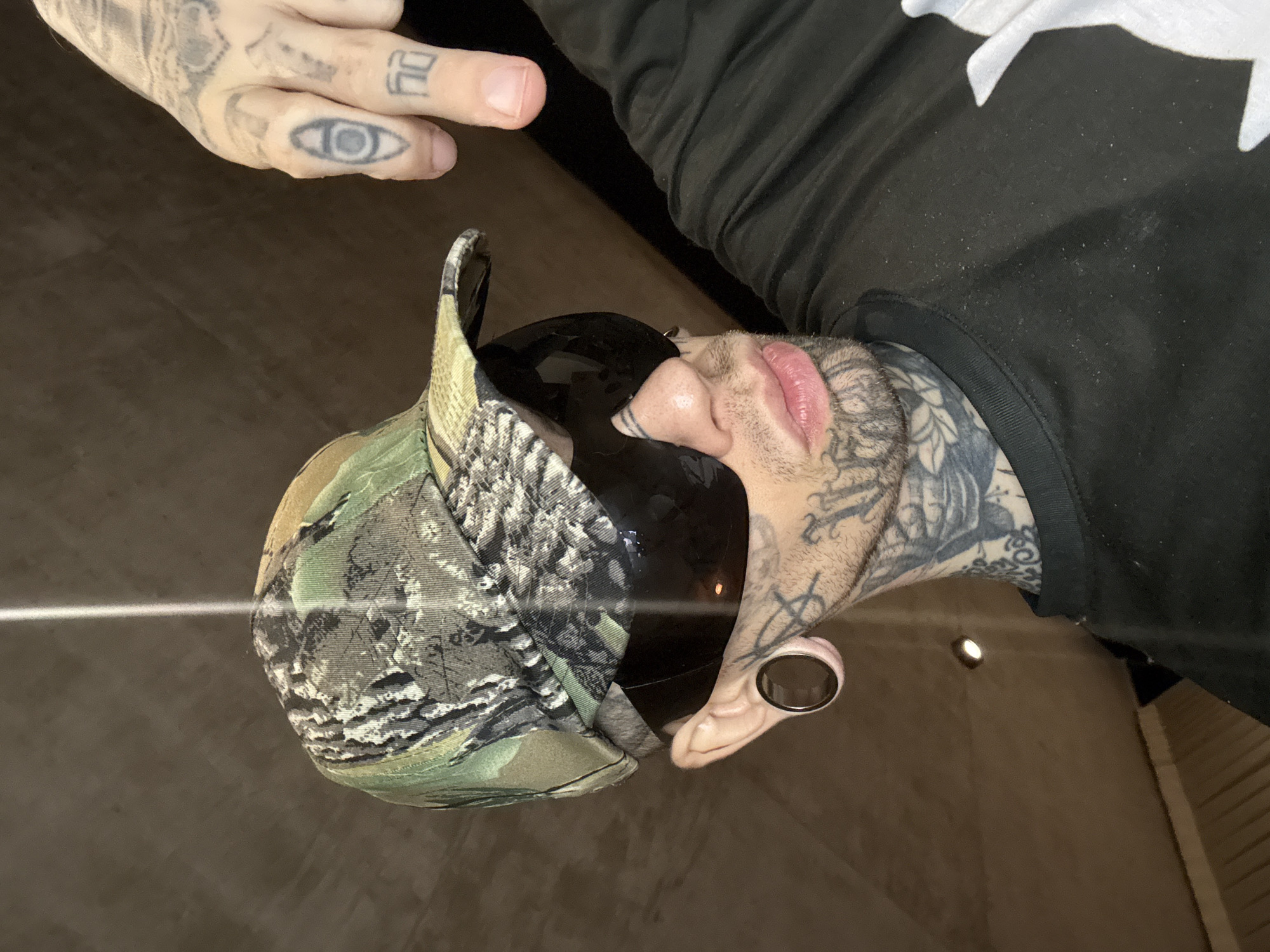 The image is a low-angle shot of a person wearing a camouflage baseball cap and dark sunglasses. The person has numerous tattoos on their face, neck, and hands, including an eye tattoo on one finger. The person is also wearing a black sweatshirt with white patterns on the upper right. The person has stretched earlobes with a large gauge earring. The person's lips are full and appear glossy. The background is blurry and suggests an indoor setting with neutral-colored walls. The person is facing towards the camera and in the city of Buenos Aires, Argentina.
