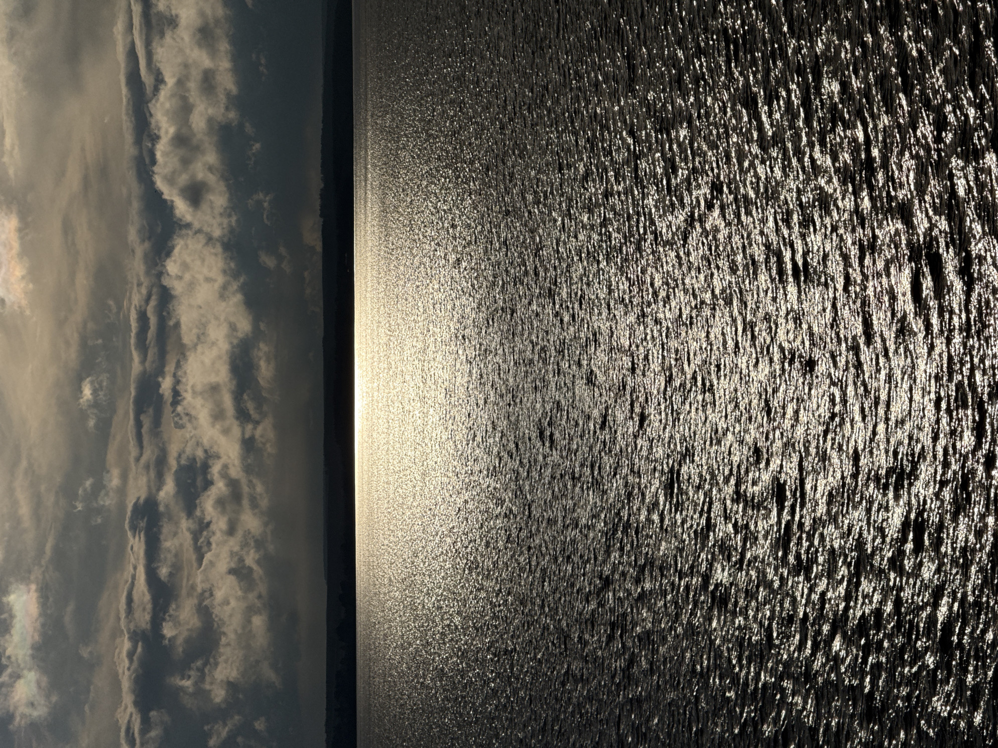 The image depicts a body of water under a sky with cloudy conditions, possibly a reservoir near Votkinsk, Russia. The water's surface is rippled and reflects sunlight, creating a sparkling effect. The sky is partially overcast, with clouds obscuring the sun to some degree. The horizon is visible in the distance.