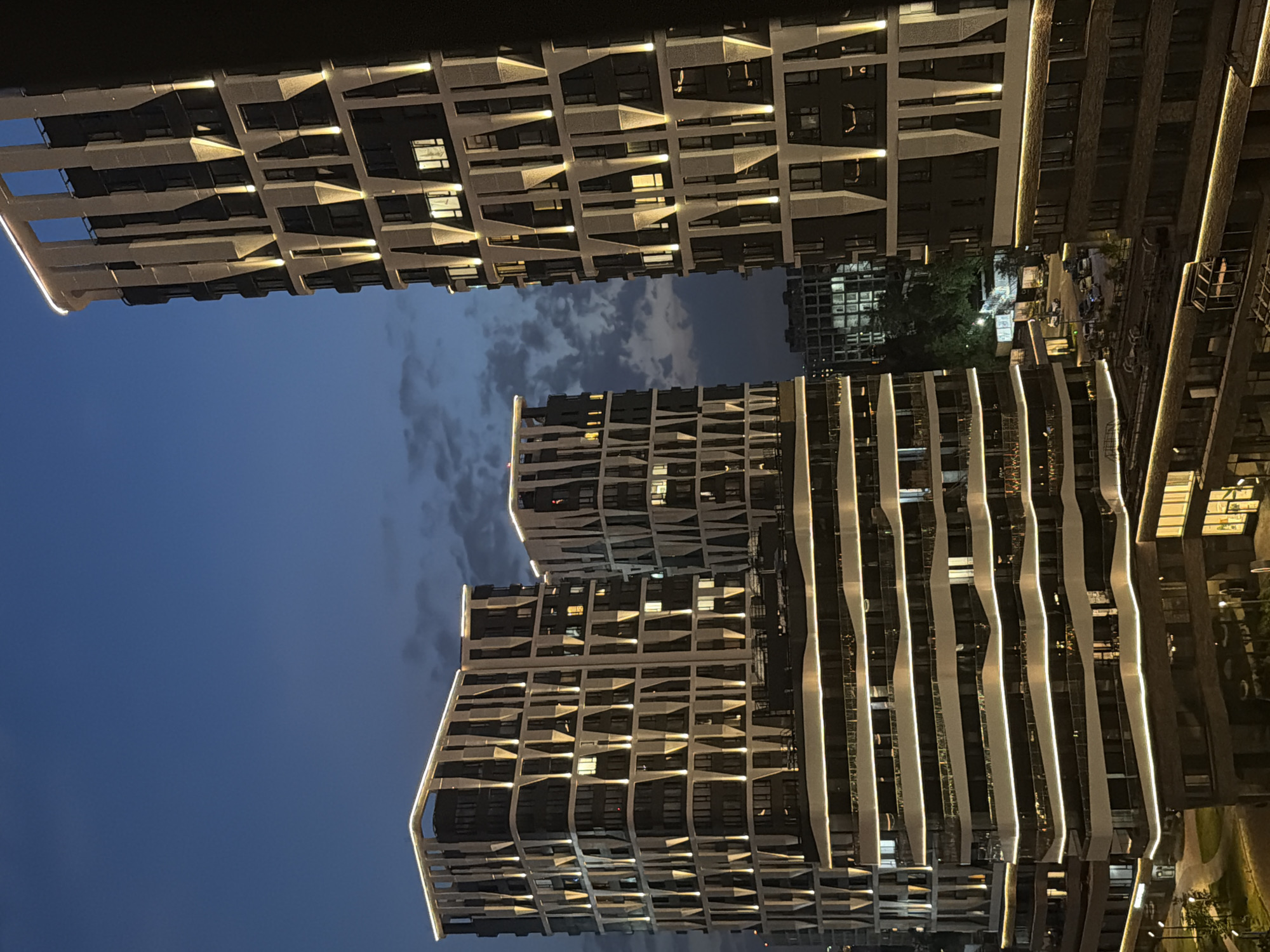 The image captures a twilight view of a modern residential complex in Moscow, Russia. The architecture is characterized by angular facades and extensive exterior lighting that outlines the building's structure and illuminates individual windows. Three prominent high-rise buildings are visible, with the one on the right being the tallest. In the foreground, lower sections of buildings with balconies and more horizontal lighting are visible, suggesting a tiered construction. The sky above is a deep blue, with faint clouds, indicating dusk or early evening. Below the residential buildings, streetlights illuminate a road with several parked cars. Trees and other structures are present in the background, suggesting an urban environment. The overall scene depicts a well-lit, contemporary urban landscape.