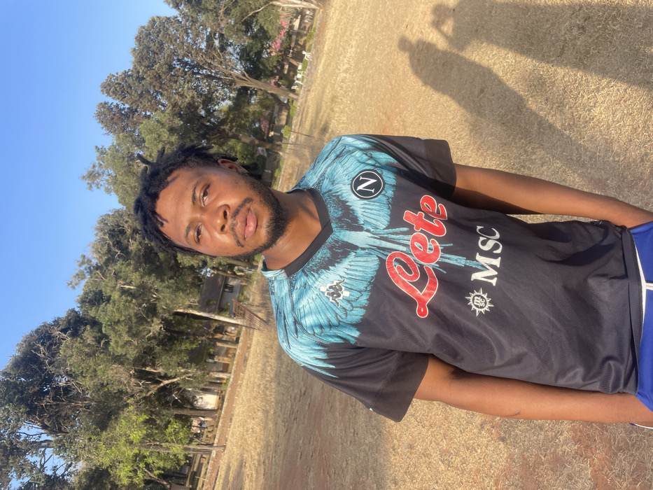 A young man with short dreadlocks and a trimmed beard stands in a grassy field. He wears a Napoli soccer jersey with blue and white wings across the shoulders, and the words "Lete" and "MSC" in red and white. The background shows trees and a clear blue sky. Sunlight casts a shadow of the person and an object in the upper right corner. The overall image is a slightly angled, medium shot.