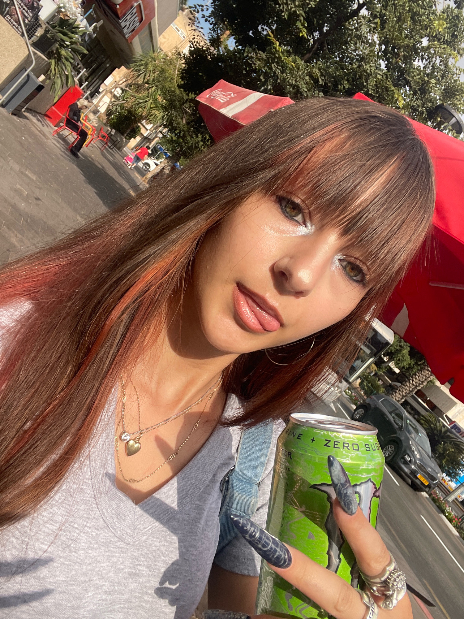 A young woman with long, brown hair, with streaks of pink, is taking a selfie in an outdoor urban setting in Raanana, Israel. She is wearing a grey t-shirt and a denim jacket, and multiple silver necklaces, one of which has a heart-shaped pendant. Her makeup includes silver eyeshadow. In her right hand, she is holding a green can of Monster Energy Zero Sugar drink, and her long, dark fingernails are painted with a grey and black marbled design and are adorned with several silver rings. She is sticking her tongue out slightly and her left eye is partially visible. In the background, there is a paved sidewalk, trees, buildings, a red umbrella, and parked cars. A person in a black suit is sitting on a red chair in the mid-ground.