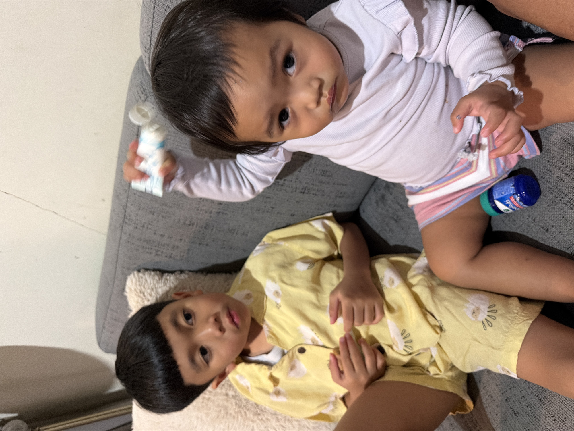 Two children are lying on a gray sofa. The younger child, an infant, is positioned above and to the right of the older child. The infant is wearing a white long-sleeved shirt and colorful striped shorts. The infant's left hand holds a small white tube with a blue cap. To the right of the infant's legs, a small blue bottle with a green cap is visible. The older child, a young boy, is wearing a yellow short-sleeved shirt with a white floral pattern. He is lying on his back, and his head is to the left of the frame. The children appear relaxed. The setting appears to be indoors, likely a living room, given the presence of the sofa. The lighting suggests it could be daytime, but there are no clear indicators of the time of day or weather. There is no visible text that can be deciphered.