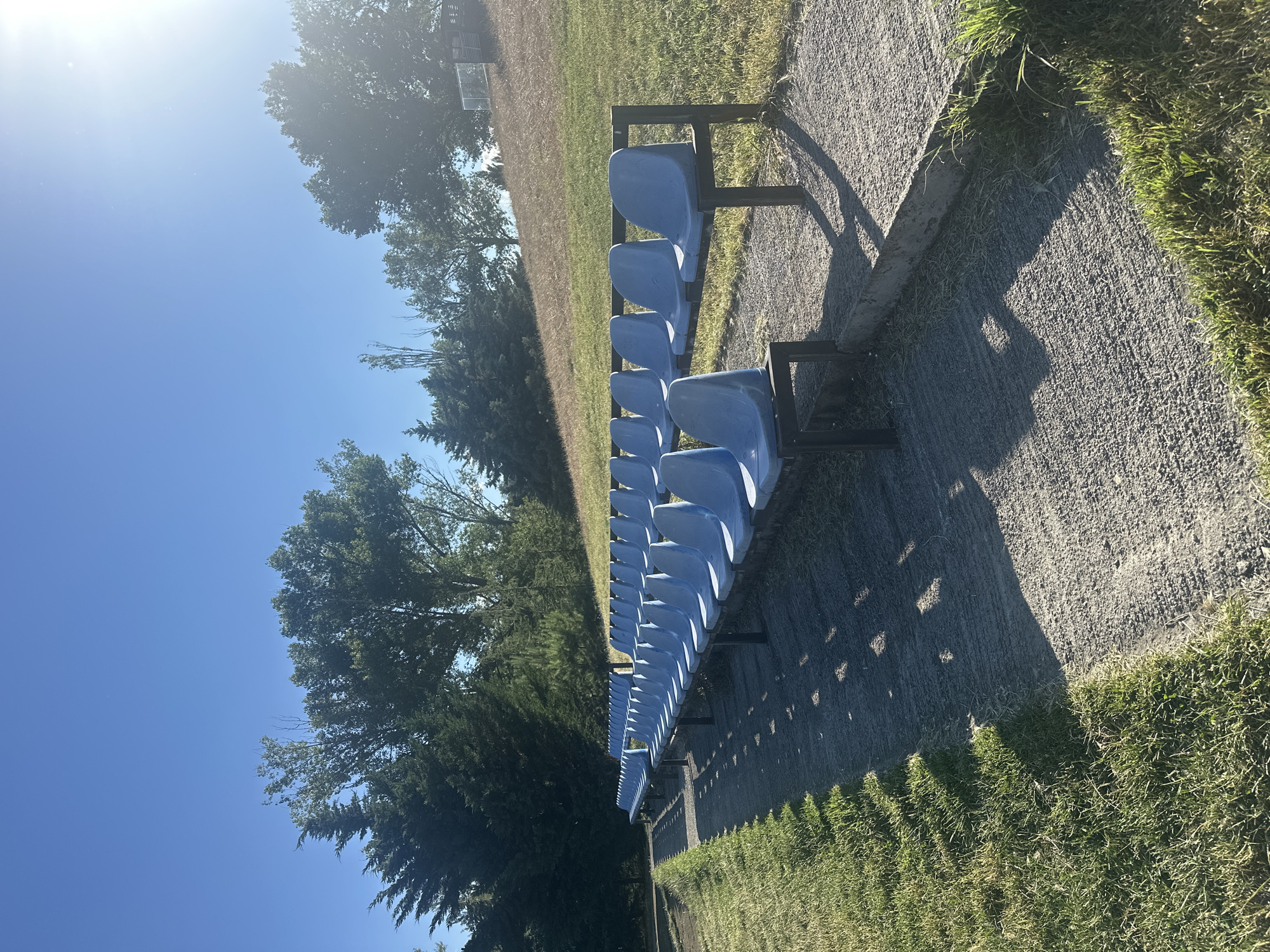 This is a rotated, medium-angle outdoor shot of bleacher seats with bright blue plastic seats mounted on black metal frames. The seats are arranged in multiple rows, facing a grassy field that extends into the background. The field is partially shaded by trees on the left side of the frame. The foreground shows a rough, concrete-like path adjacent to the bleachers, with tufts of green grass growing along its edge. Long, dappled shadows are cast across the path from the bleacher structure, suggesting it is a sunny day. The sky is a clear, vibrant blue, with the sun prominently visible in the upper left quadrant, its glare creating a bright haze. The overall scene evokes a sense of an outdoor sporting venue or recreational area, likely in a temperate climate. The time of day appears to be mid-morning or afternoon, given the angle and length of the shadows and the bright sunlight. No people are visible in the image. The location is identified as Tbilisi, Georgia.